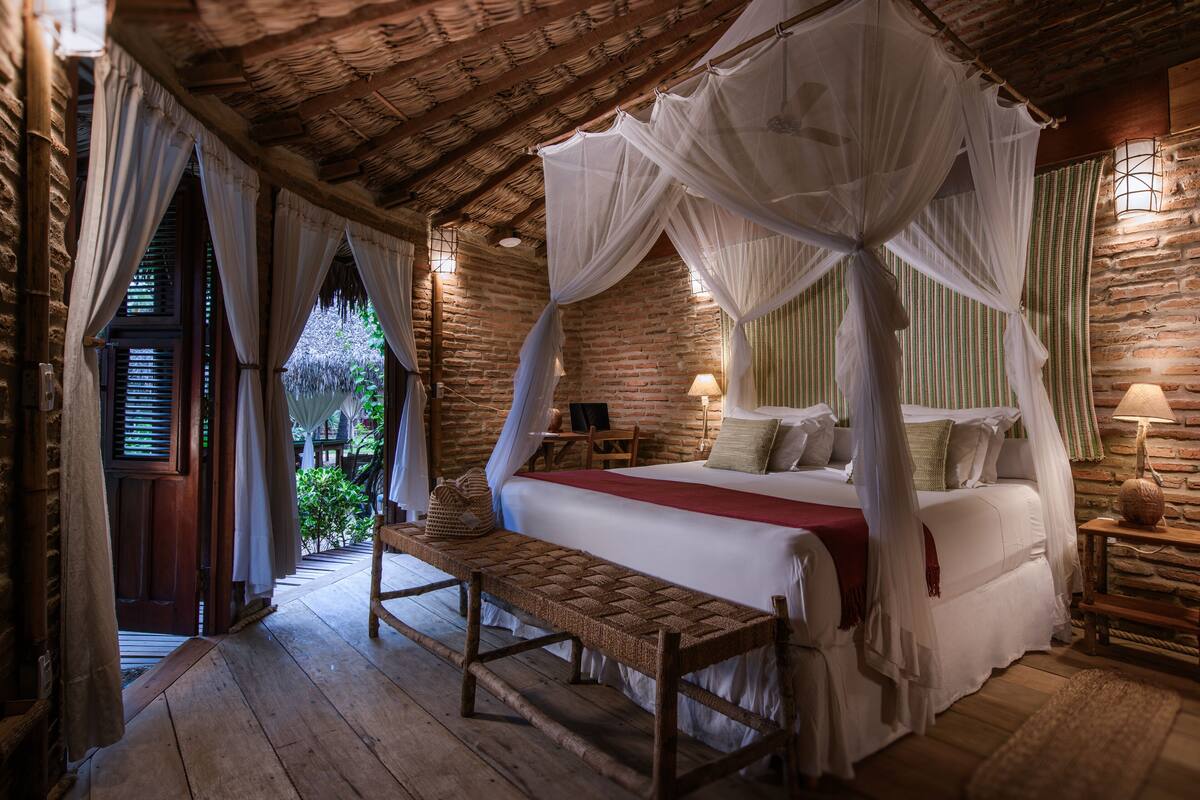 A room with a bed under a canopy, a bench, a lamp, and a view of greenery outside.