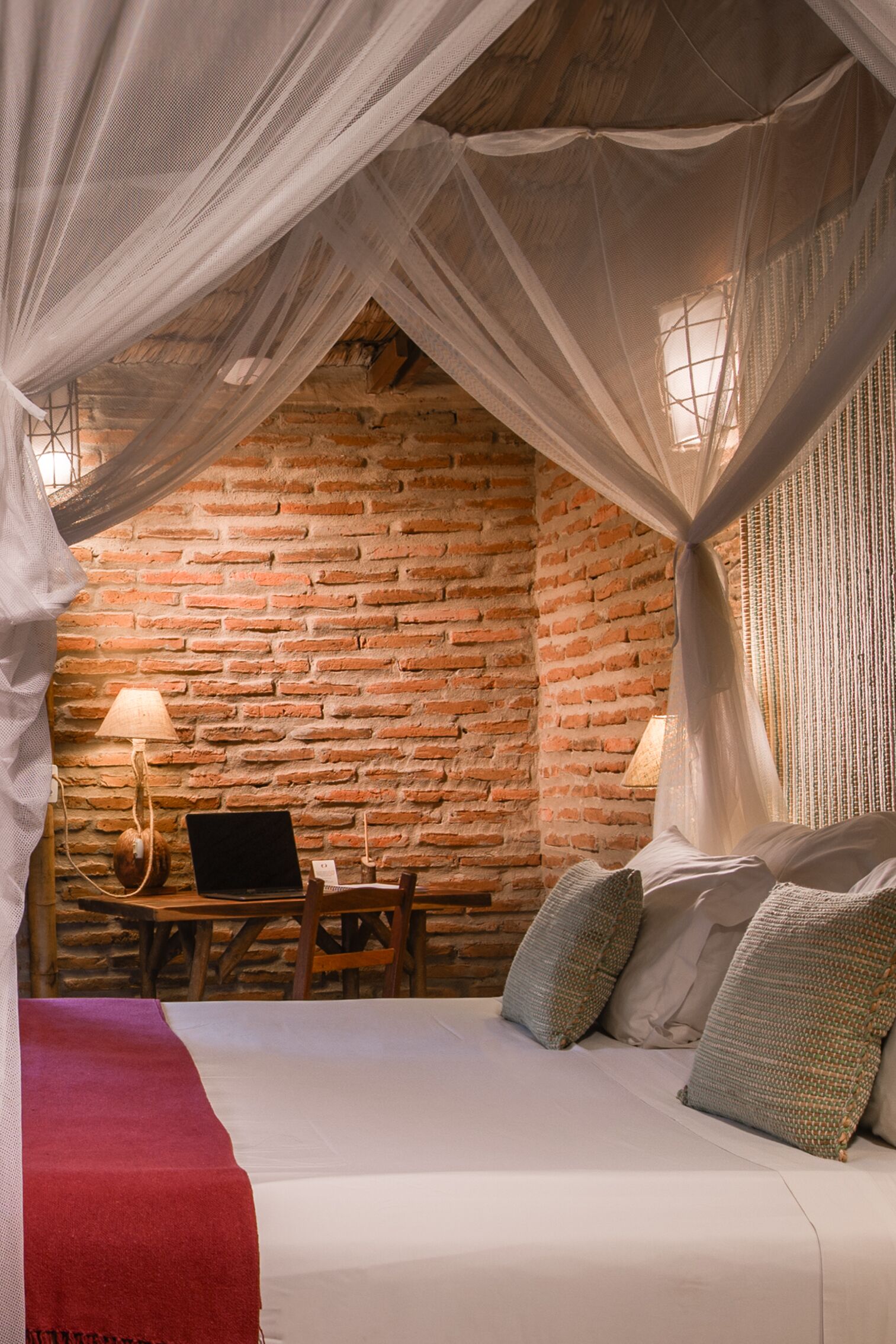 A room with a bed under a canopy, a desk with a laptop, and a brick wall.