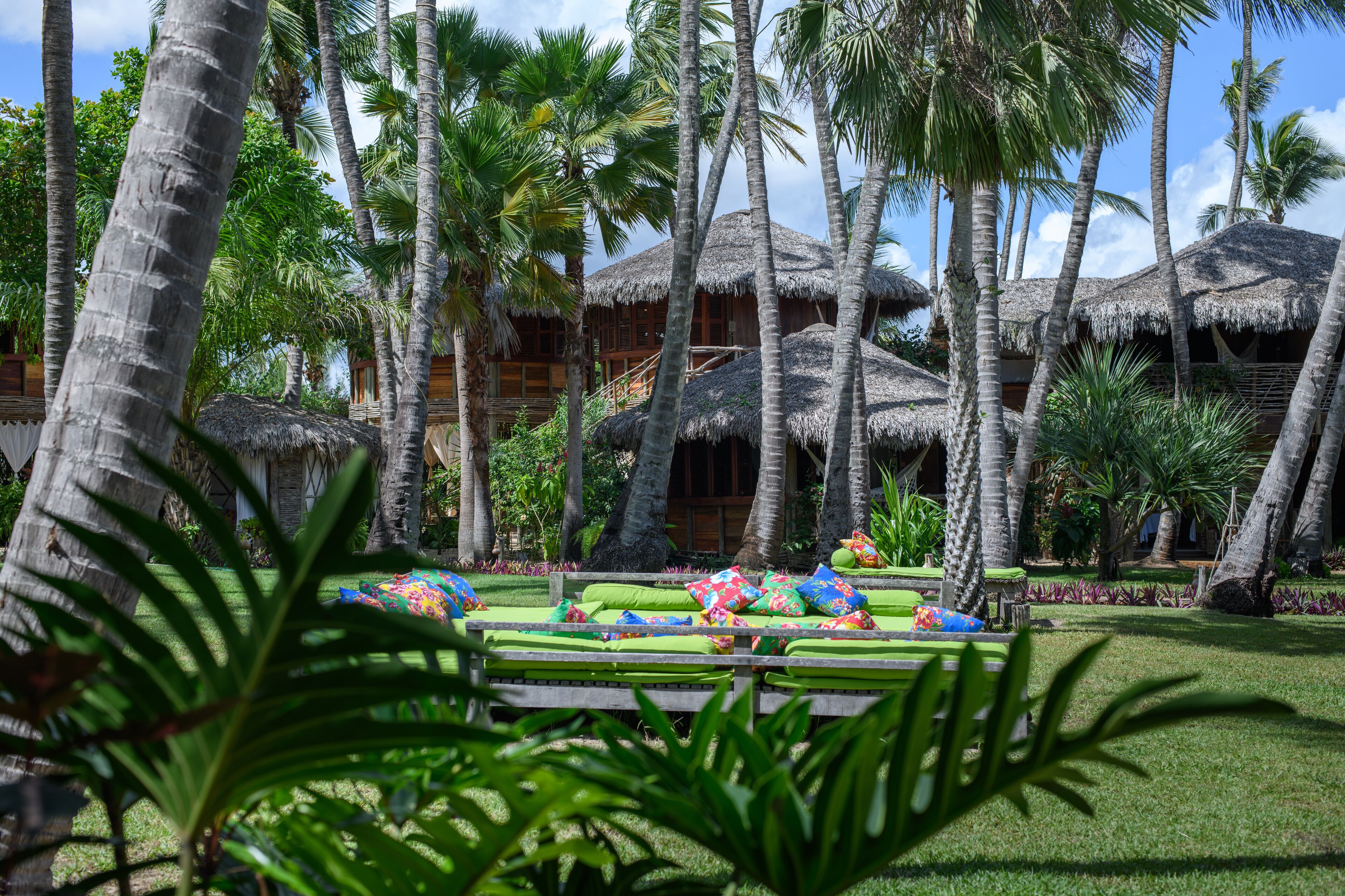 Un complexe hôtelier doté de bungalows aux toits de chaume, de palmiers et d’espaces de détente en plein air agrémentés de coussins colorés.