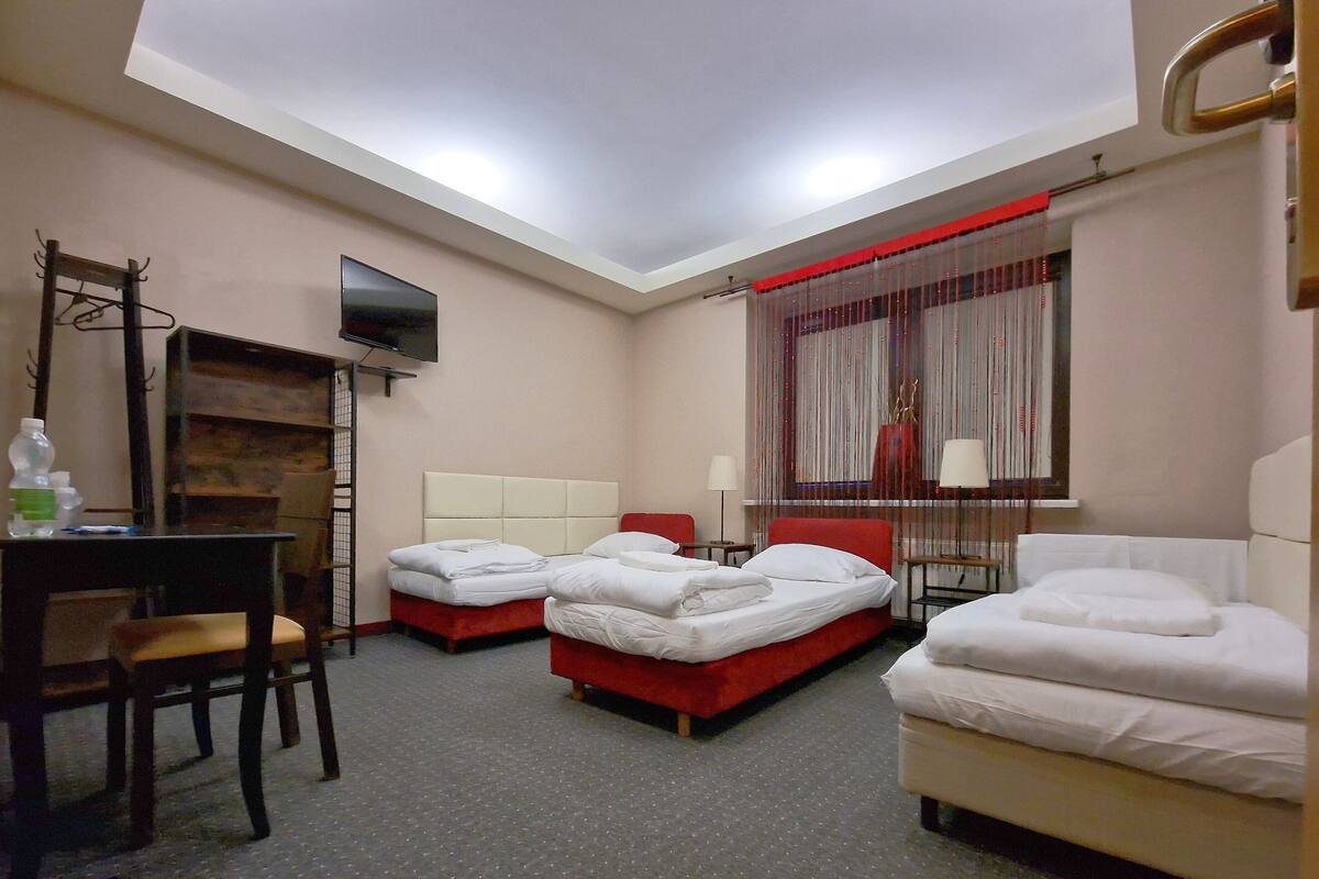 A hotel room with three beds, a desk, a chair, a TV, and a window with curtains.