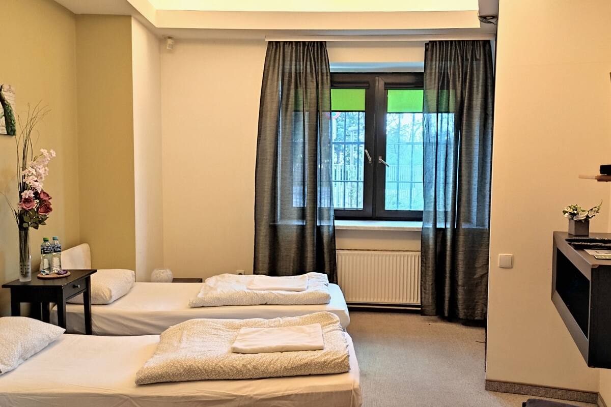 A hotel room with two beds, a desk with a TV, a radiator, and a window with curtains.