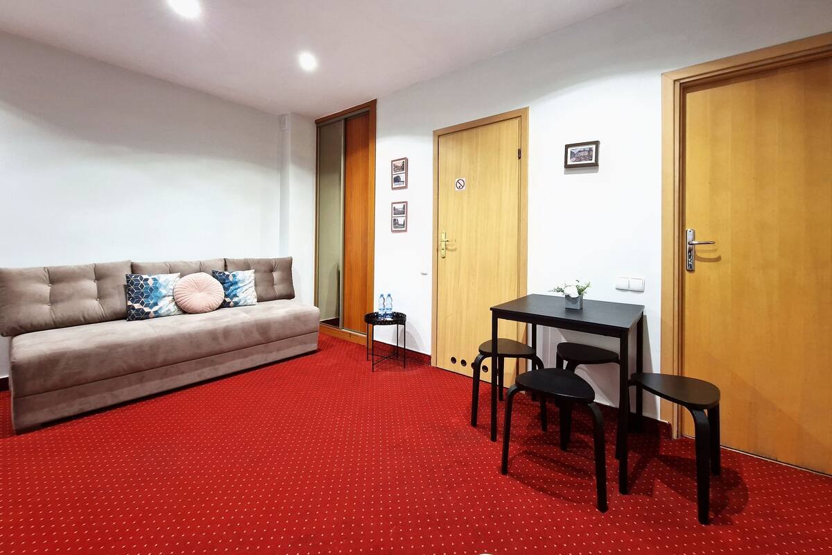 A room with a sofa, a small table with chairs, a red carpet, and wooden doors.