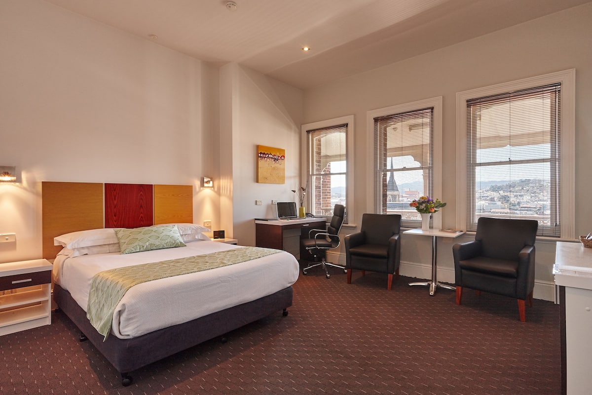 Habitación de hotel con cama, escritorio, dos sillas, una mesita y ventana con persianas.