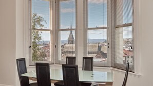 A dining area with a glass table and black chairs, large windows with blinds, and a view of a cityscape including a church spire. - Auldington Hotel (Launceston)