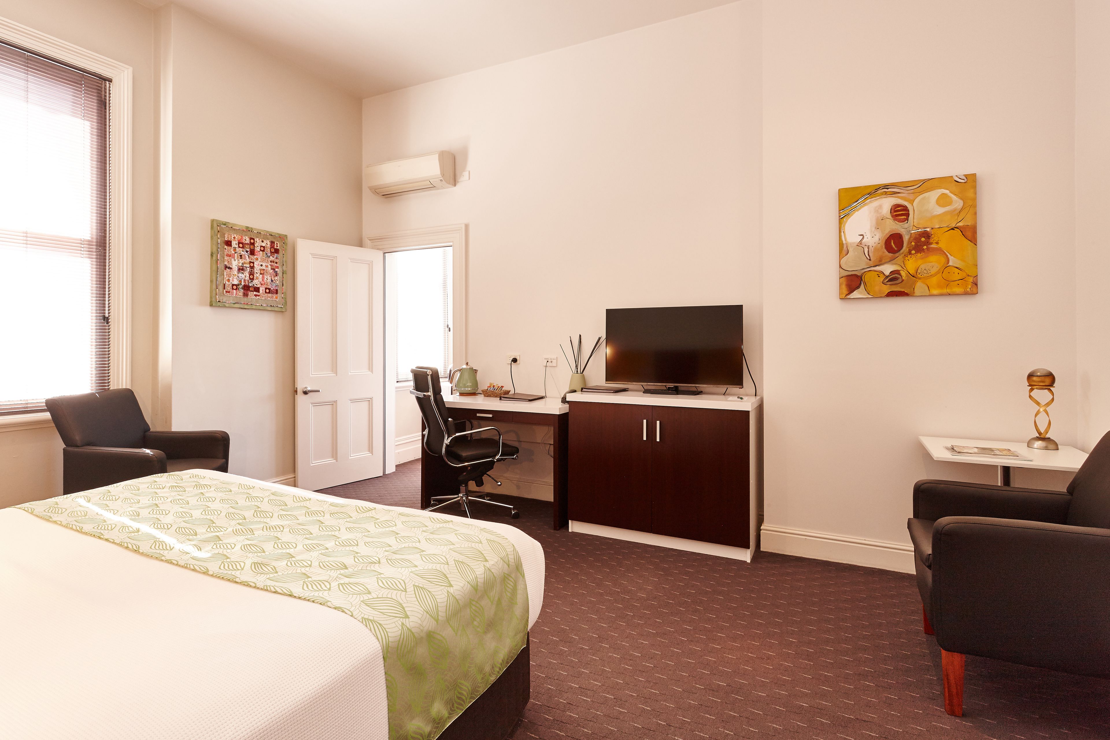 A hotel room with a bed, desk, TV, two chairs, and artwork on the wall.
