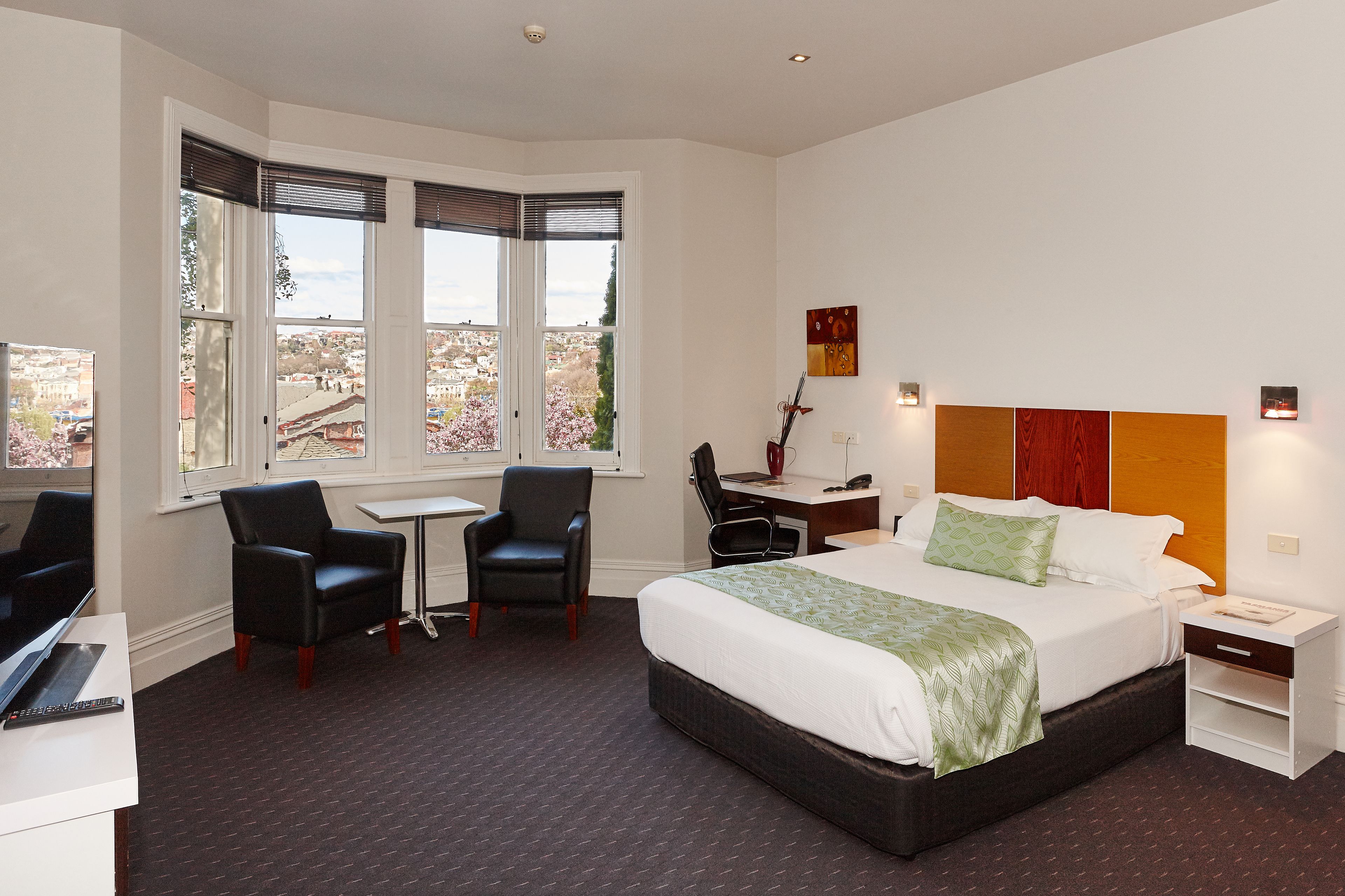 Ein Hotelzimmer mit Bett, Schreibtisch, Stuhl und Blick auf die Stadt durch große Fenster.