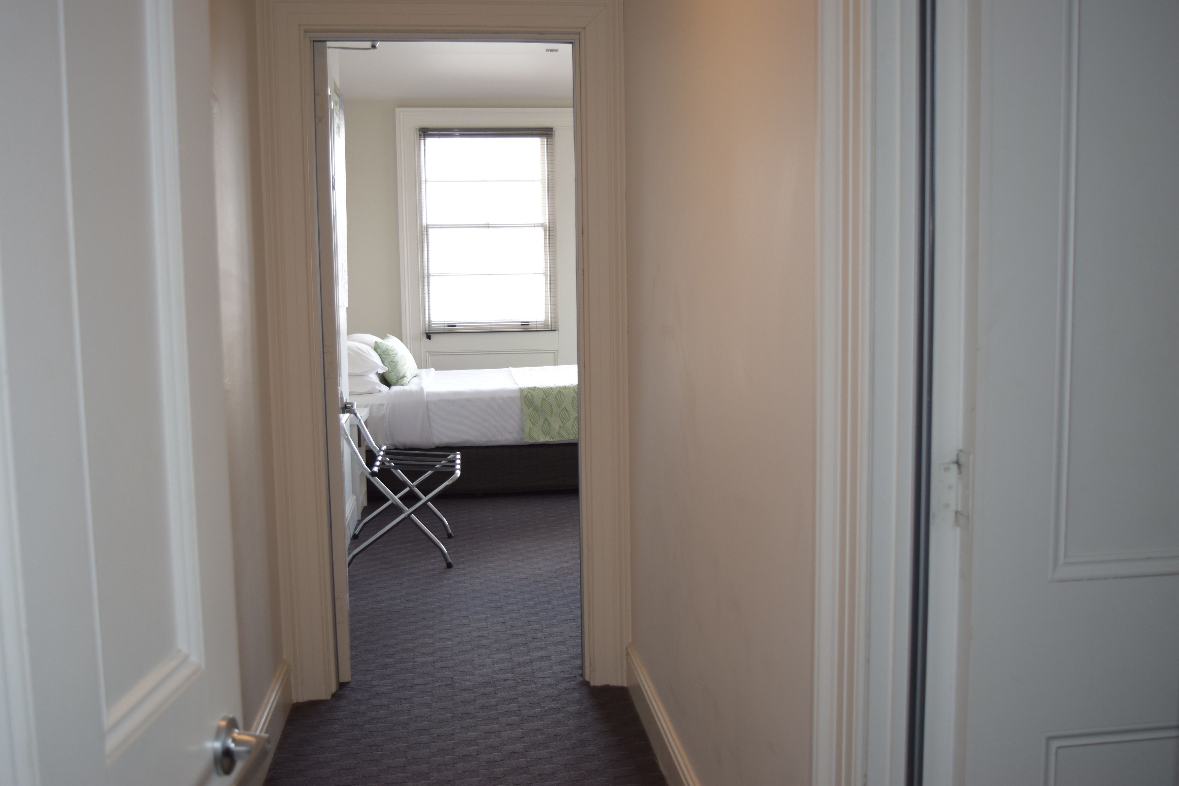 A narrow hallway leading to a bedroom with a bed, a window, and a drying rack.