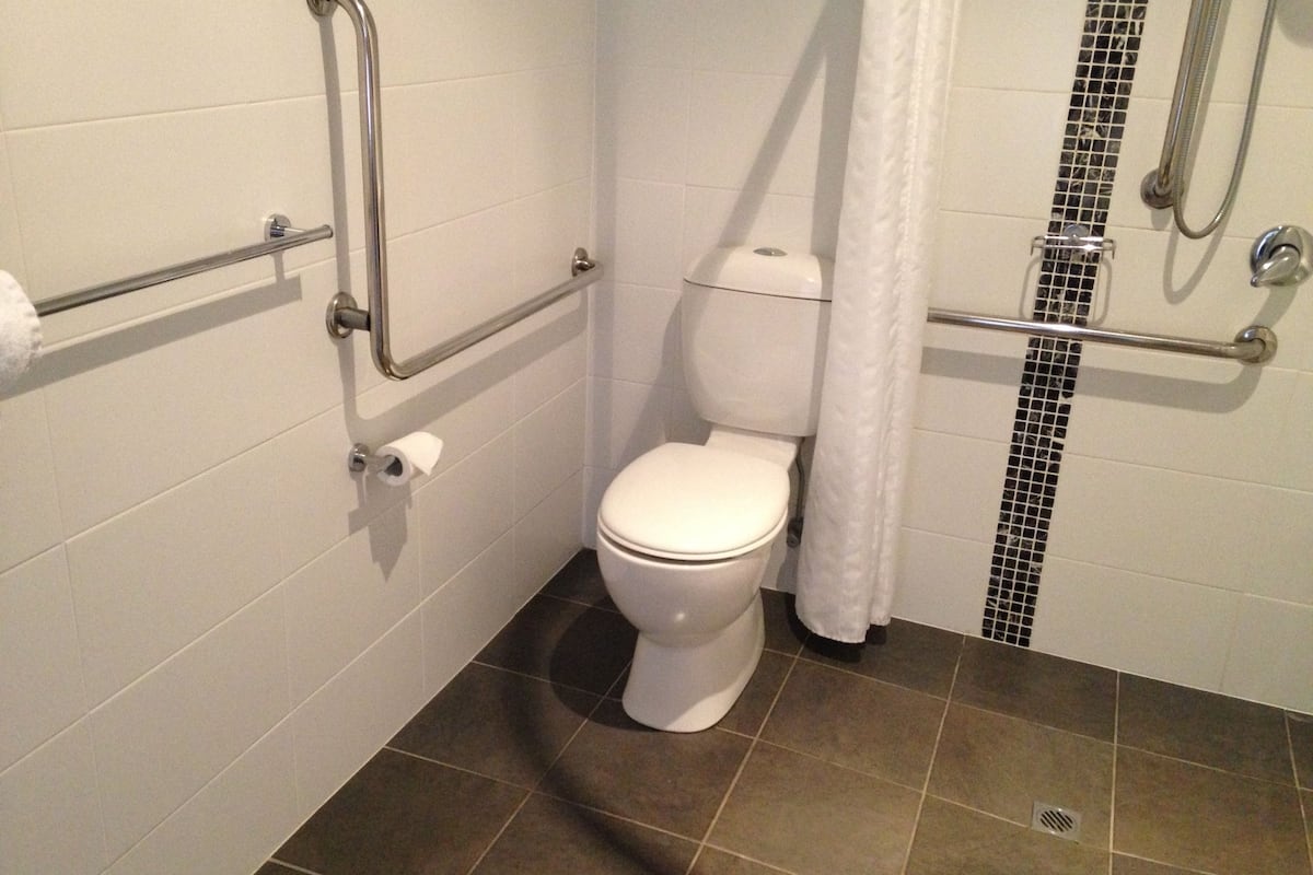 Un baño con inodoro blanco, barras de apoyo, zona de ducha y suelo de baldosas.