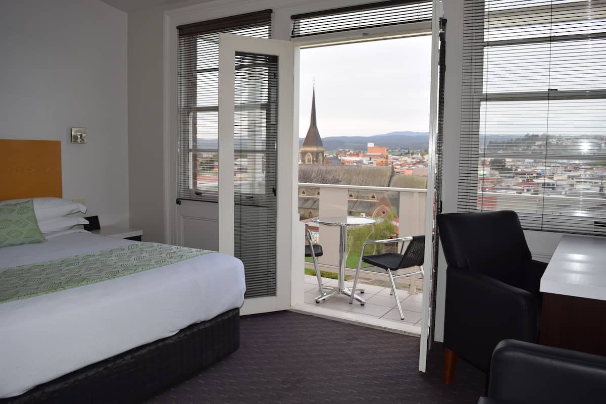 Habitación de hotel con cama, escritorio y silla. La habitación tiene vista a un paisaje urbano con una aguja de iglesia.