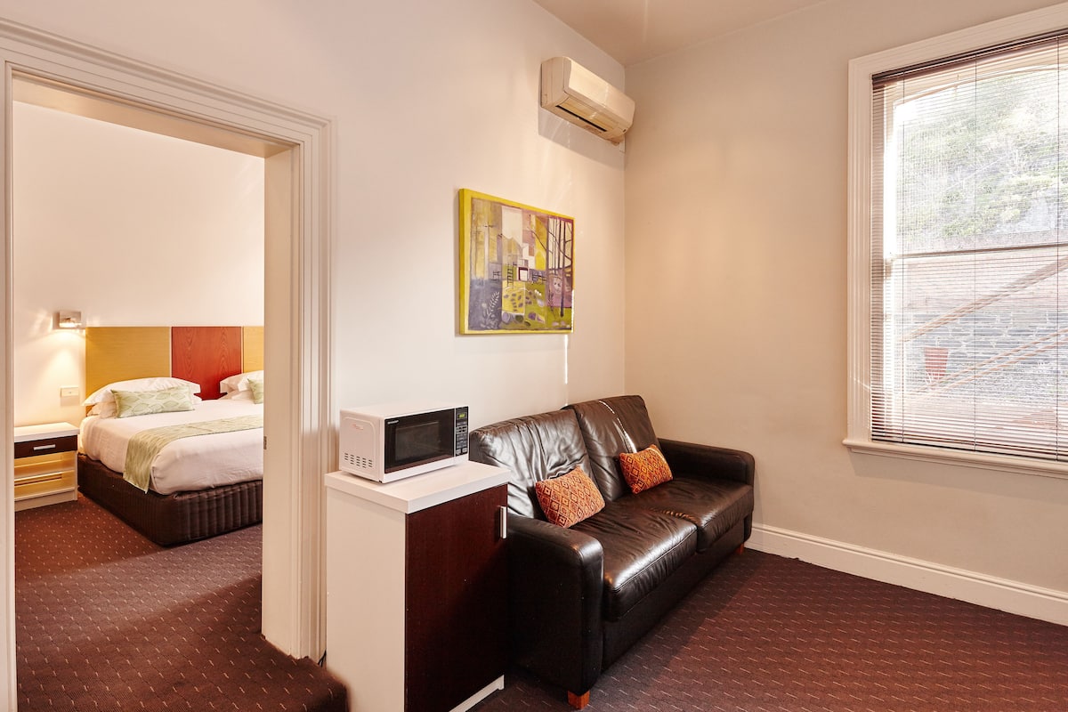 Una habitación de hotel con una cama, un escritorio con microondas, un sofá de cuero y una ventana con persianas.