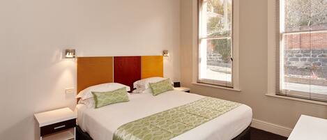 Una habitación de hotel con una cama, mesitas de noche y dos ventanas con persianas.