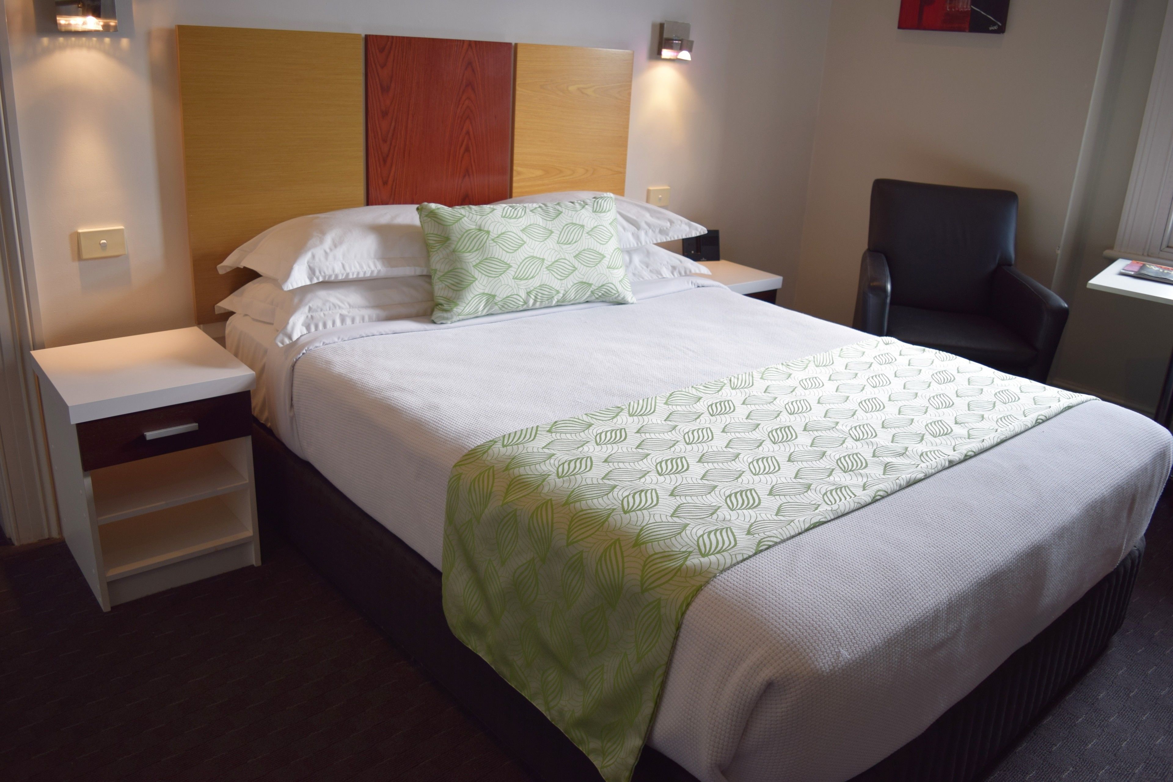 A neatly made bed with a patterned bedspread and pillows, a wooden headboard, bedside tables, and a chair.