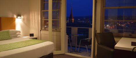 Habitación de hotel con cama, escritorio y silla. Se ve una ventana con vista a la ciudad y la aguja de una iglesia.