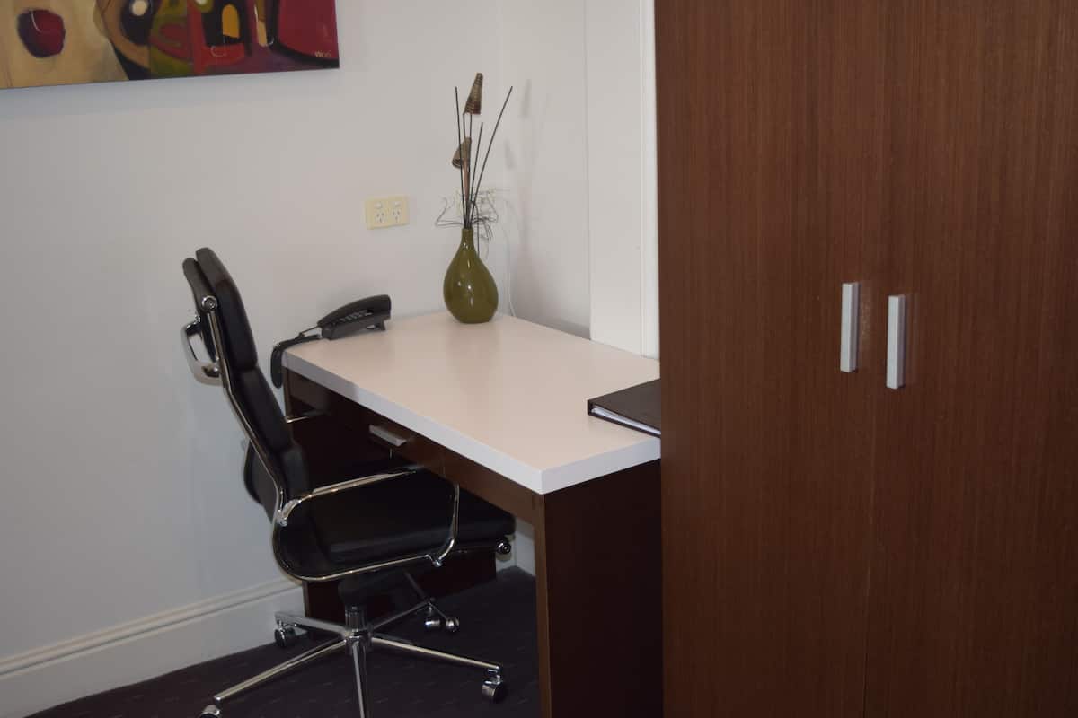 Un pequeño espacio de oficina con un escritorio, una silla de oficina negra, un teléfono y un florero con plantas secas.