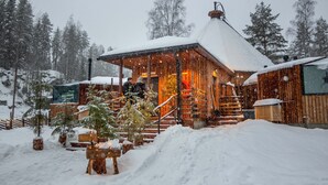 A wooden cabin with a snow-covered roof and a lit interior, surrounded by snow and trees.