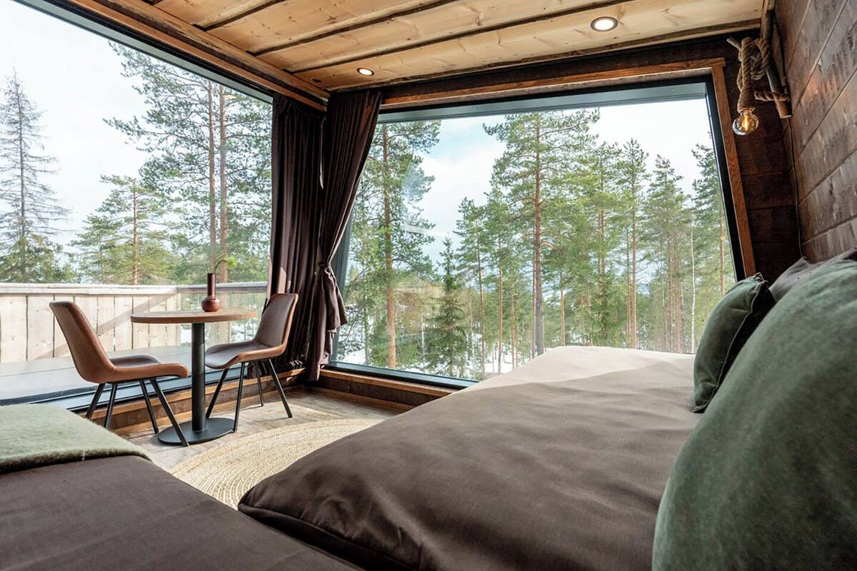 Ein Schlafzimmer mit einem großen Bett, einem kleinen Tisch, zwei Stühlen und Blick auf Bäume durch ein großes Fenster.