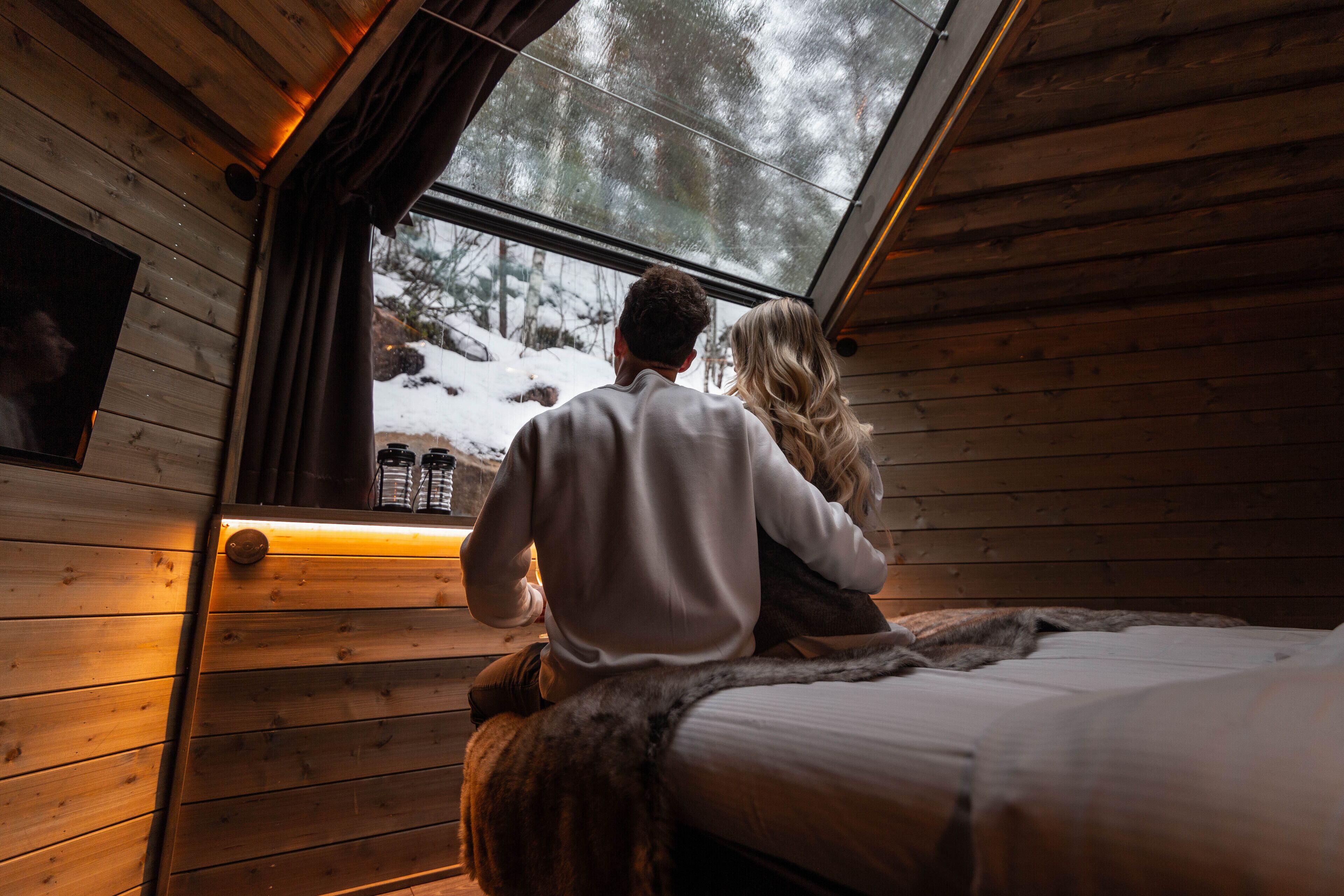 A couple sitting on a bed in a cabin with wooden walls and a large window overlooking a snowy landscape.