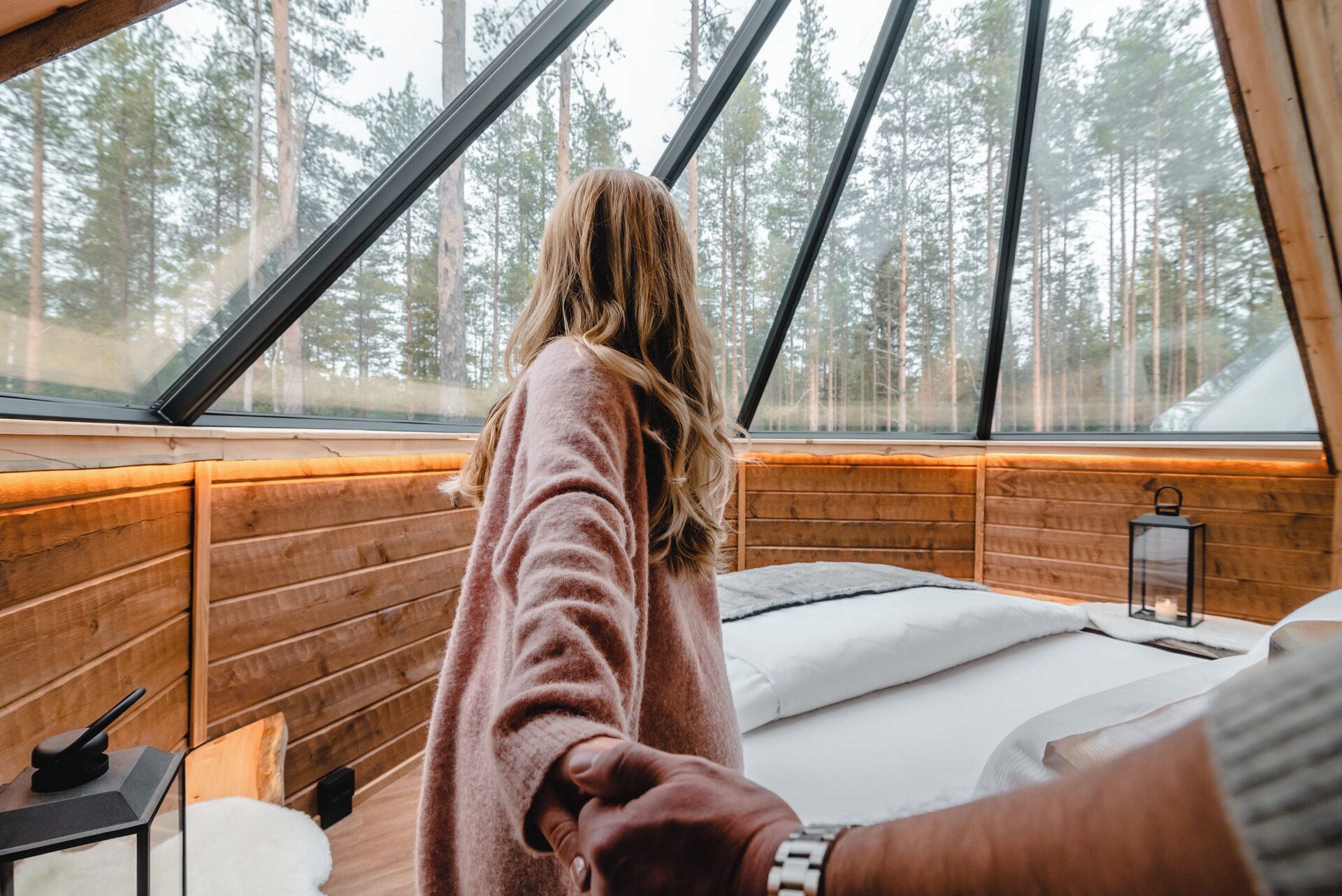 A cozy cabin interior with a large window overlooking a forest, wooden walls, a bed with white bedding, and a small table with a lantern.