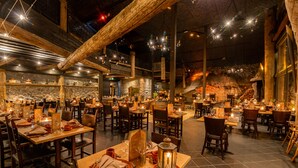 A restaurant with multiple dining tables and chairs, wooden beams, stone walls, and hanging lights.