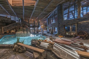 Amplia zona de piscina interior con terraza de madera, sillas de descanso y una cascada de piedra.