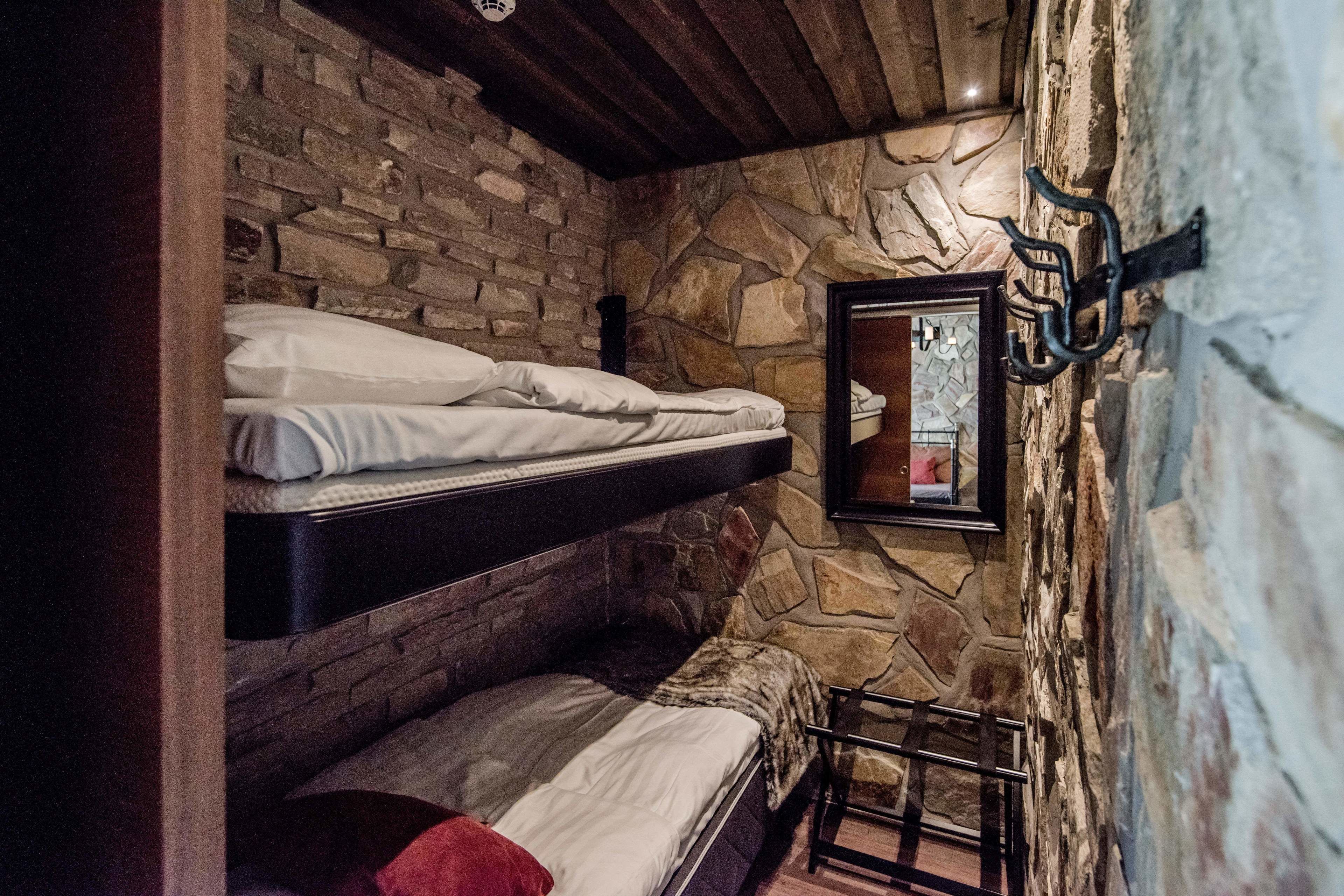 A room with a stone wall and wooden beams, featuring two bunk beds and a small table with a chair.