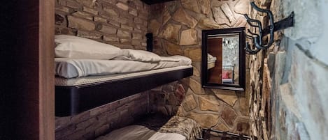 A room with a stone wall and wooden beams, featuring two bunk beds and a small table with a chair.