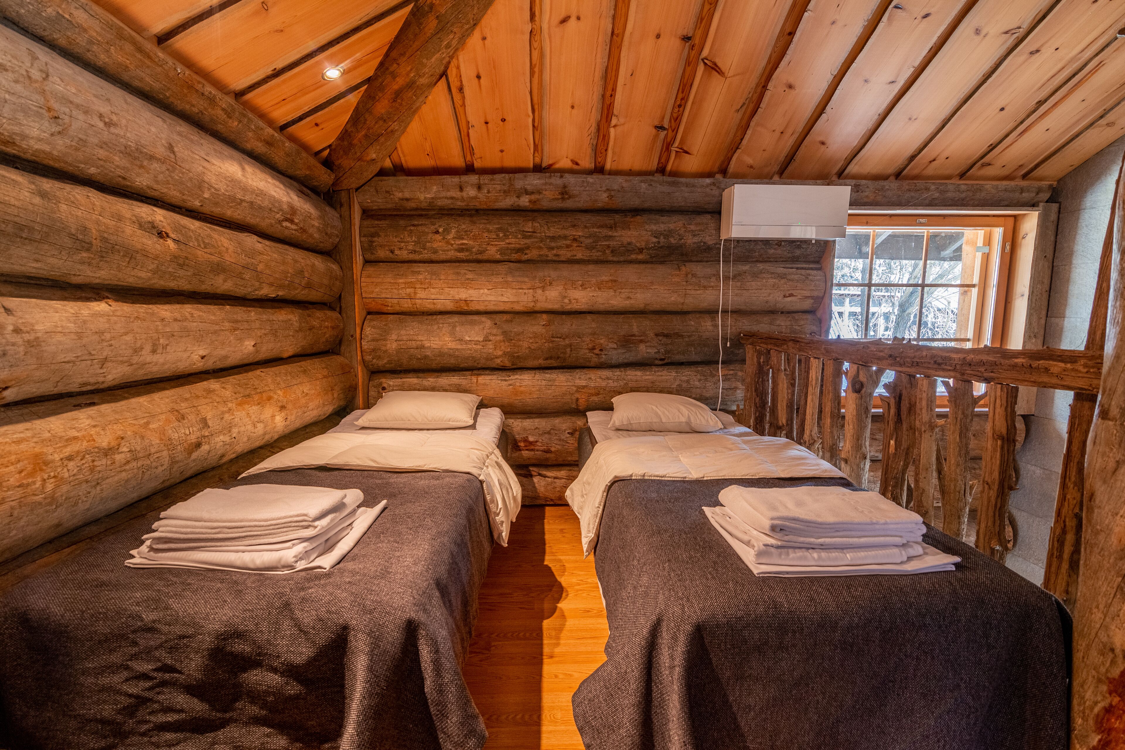 A log cabin room with two beds, wooden walls, a window, and a balcony.