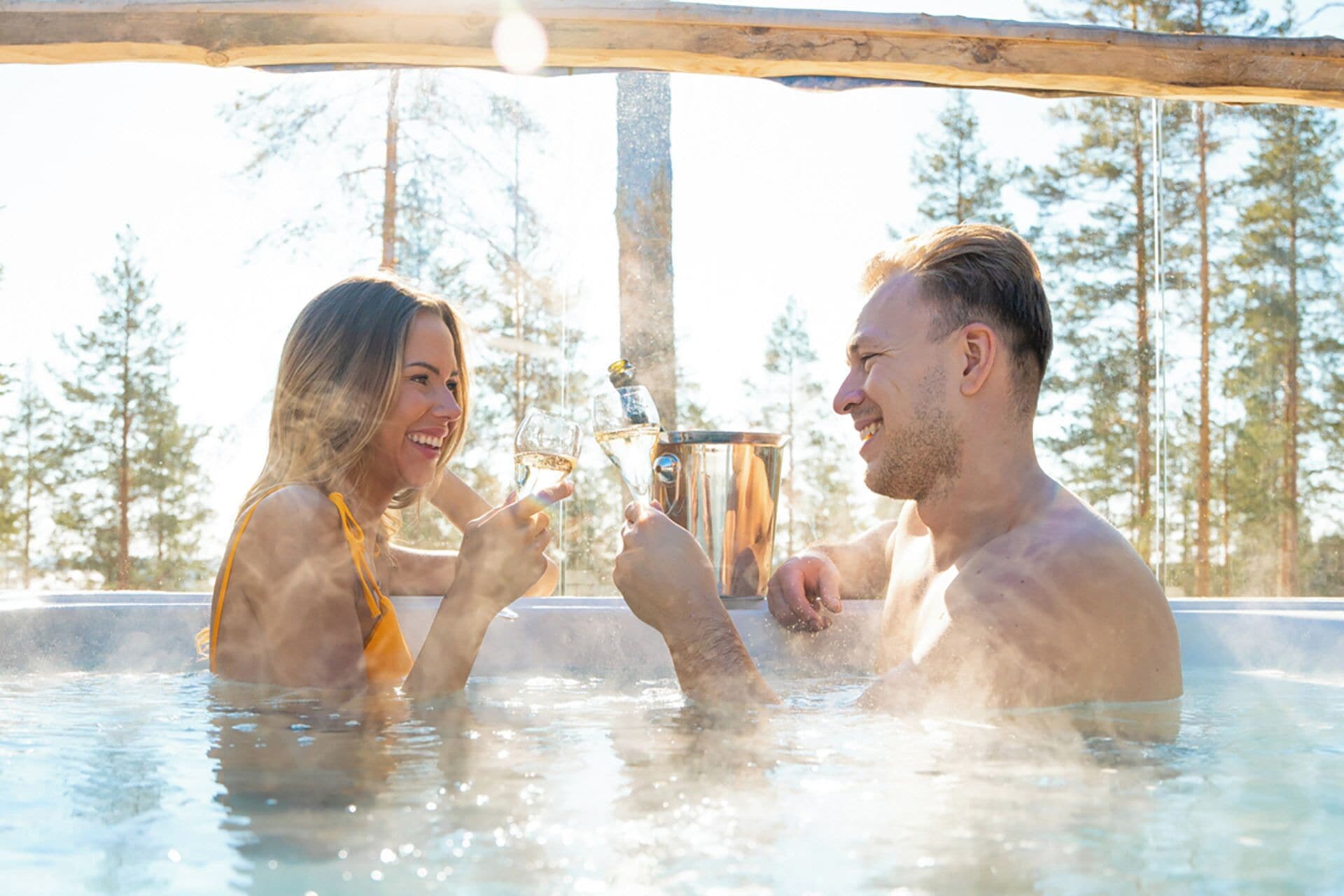 Twee personen in een jacuzzi, overdekt met een houten constructie, omgeven door bomen.