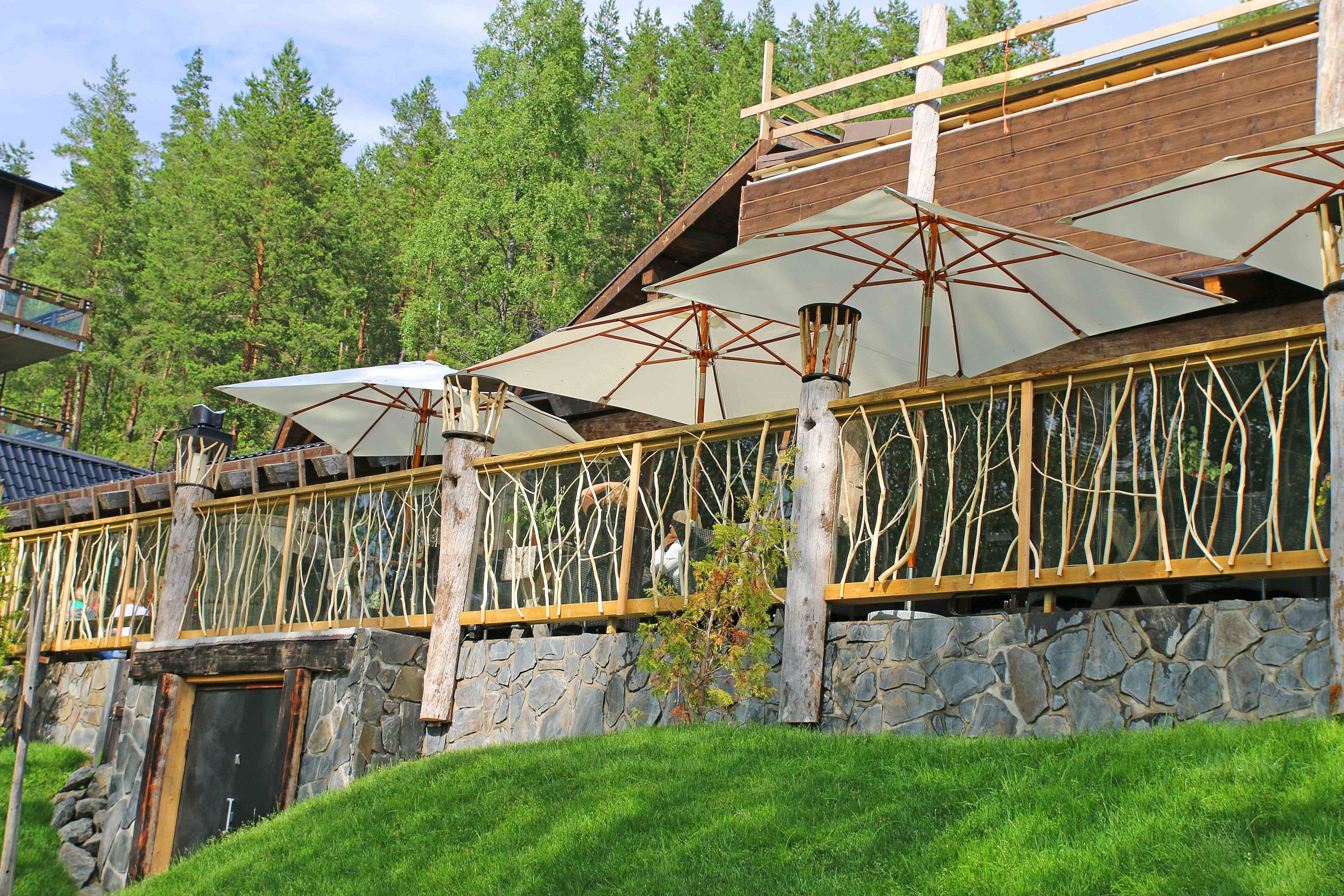 A rustic wooden building with stone walls, large umbrellas, and a wooden railing overlooking a forested area.