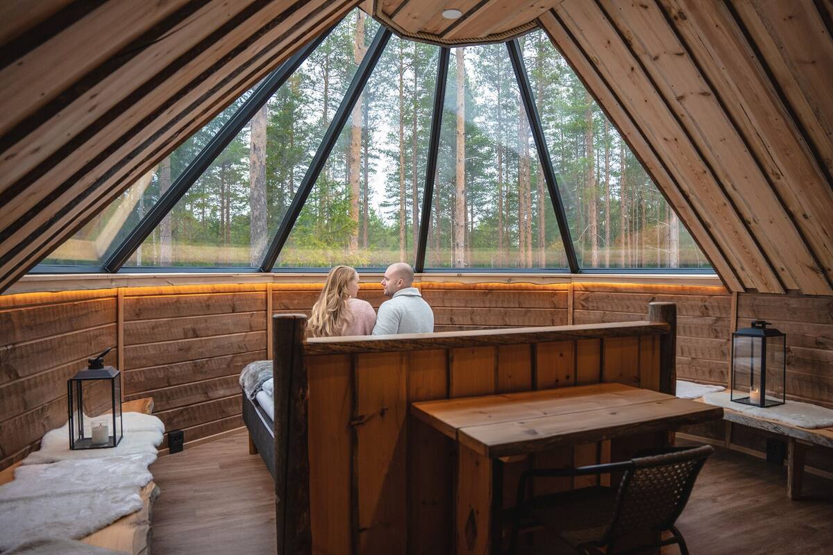 Ein gemütliches Blockhaus mit Holzvertäfelung, einer Bank, einem Tisch und einem großen Fenster mit Blick auf einen Wald.