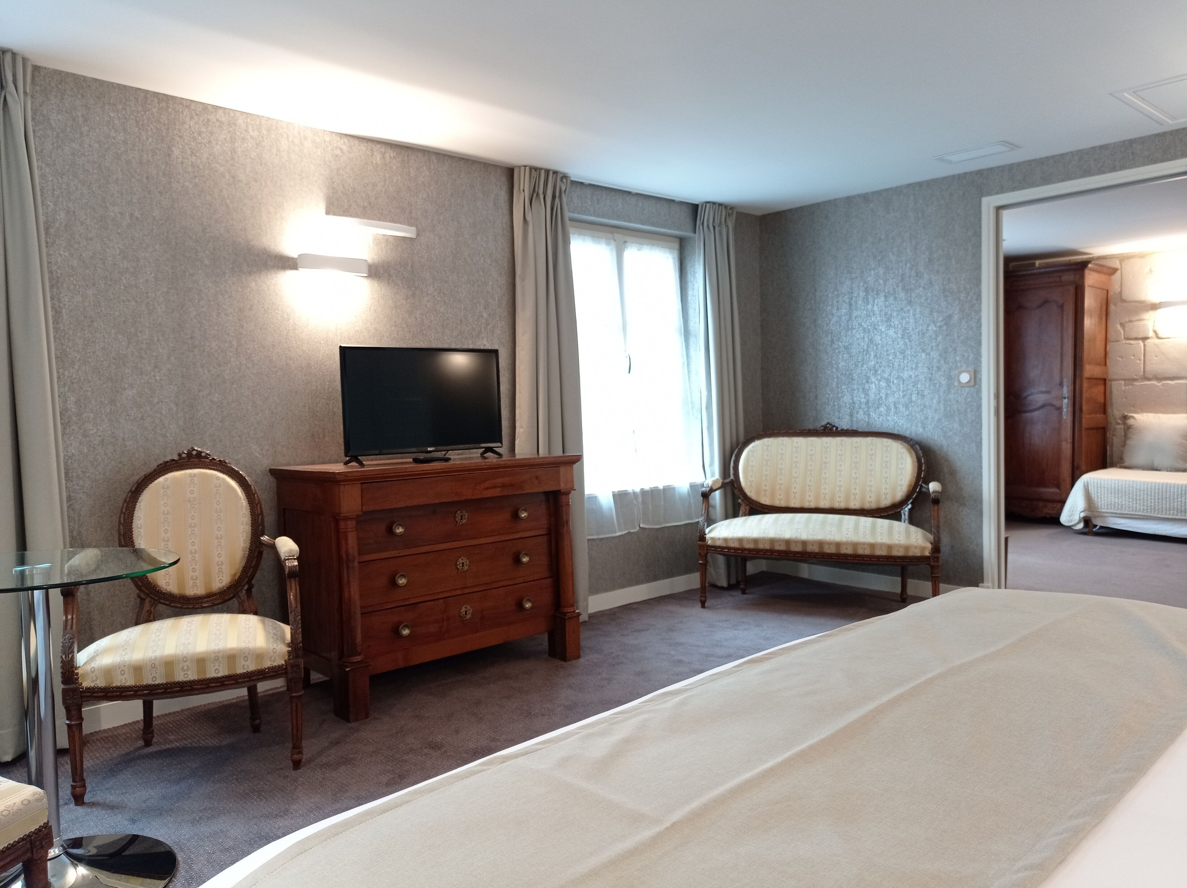 A hotel room with a bed, a wooden dresser, a television, two upholstered chairs, and a glass side table.
