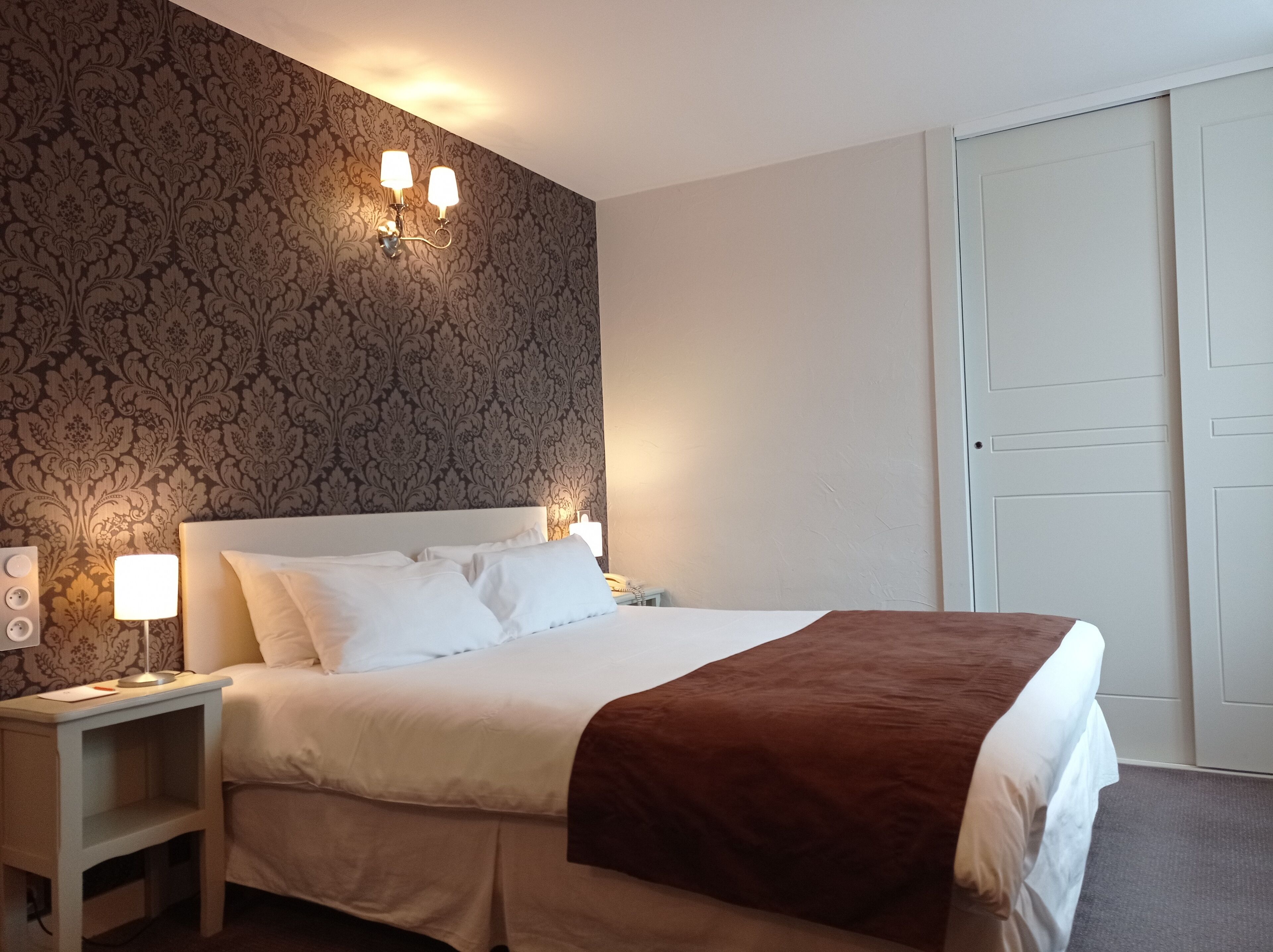 Una cama bien hecha con sábanas blancas y un plaid marrón, una lámpara de noche, y una pared con estampado floral.