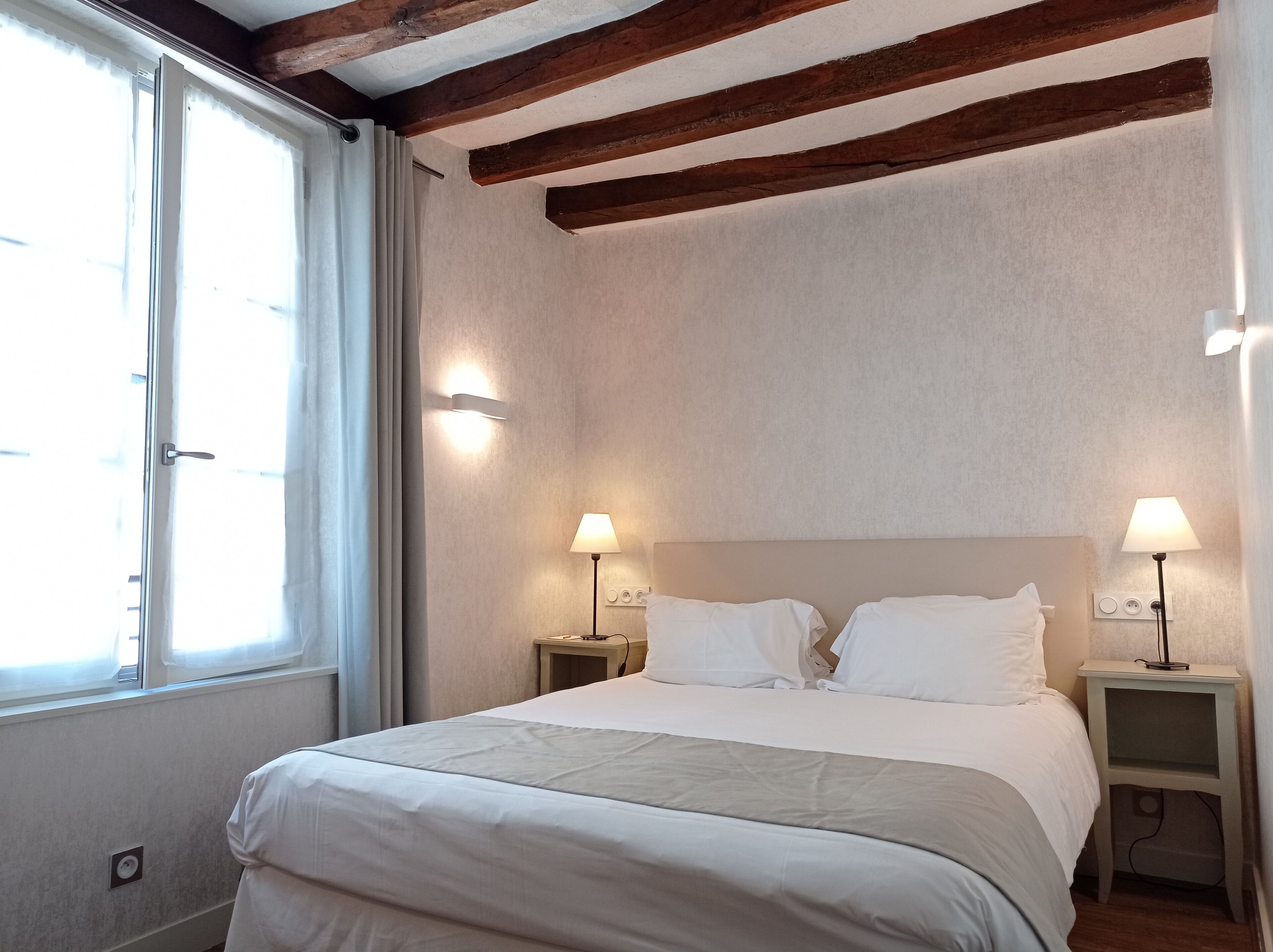Een slaapkamer met een groot bed, twee nachtkastjes met lampen, een raam met gordijnen, en zichtbare houten balken aan het plafond.