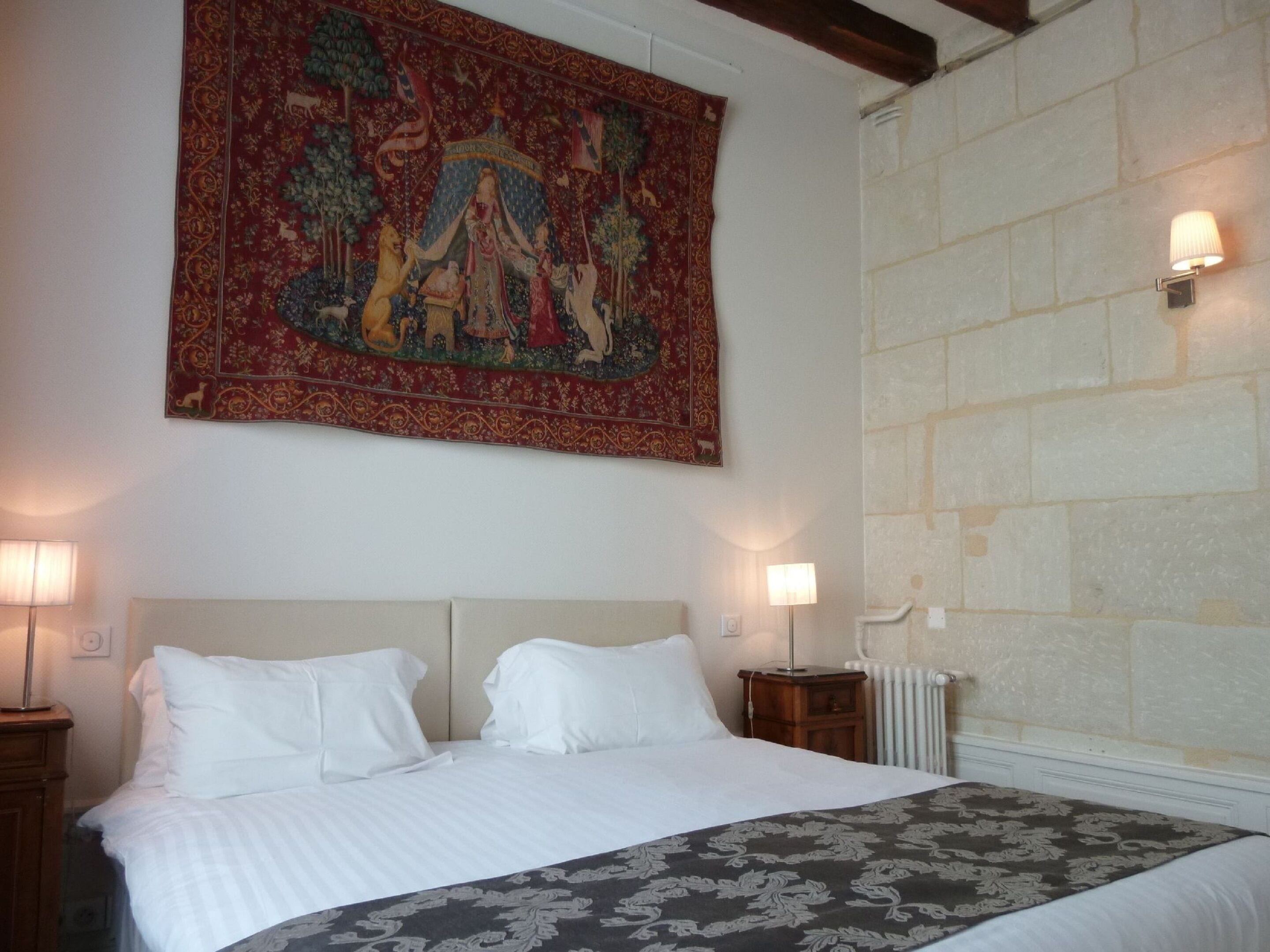 A hotel room with a large tapestry, a bed with white linens, two bedside lamps, and a stone wall.