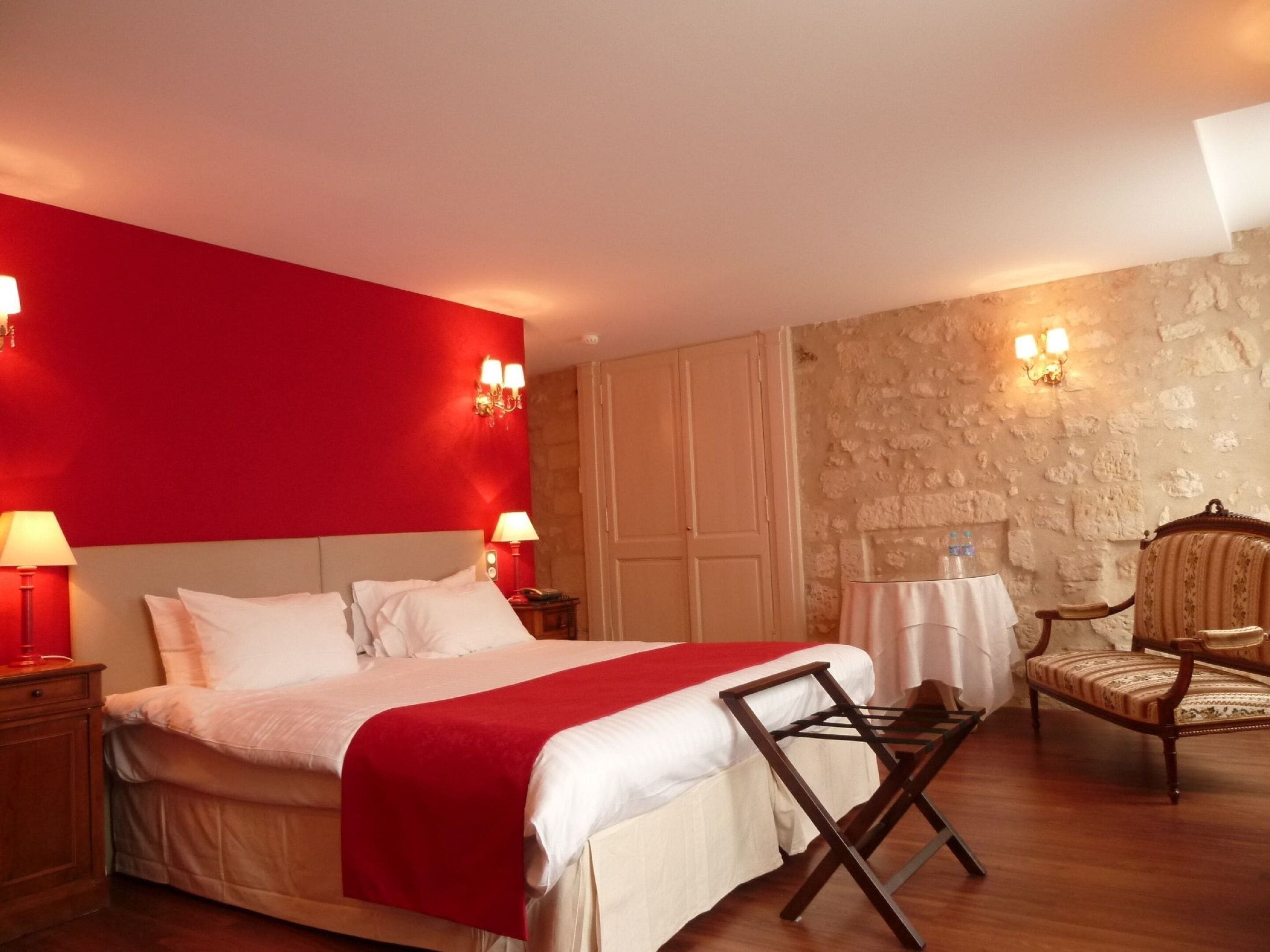 Habitación de hotel con una cama grande, una silla de madera, una mesa con una botella de agua y una pared de piedra.