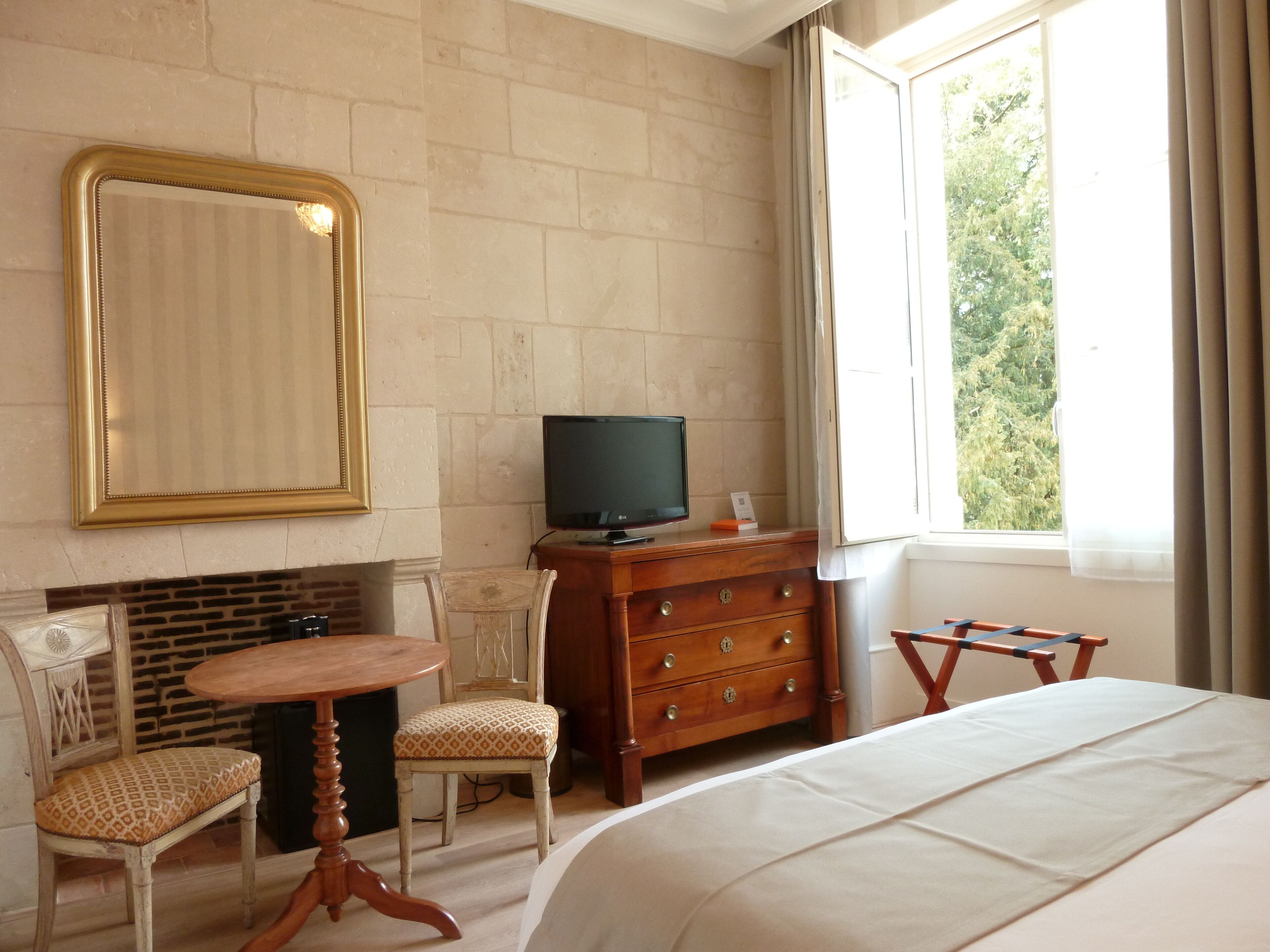 A hotel room with a bed, a television on a wooden dresser, a small table with a chair, and a large window with curtains.