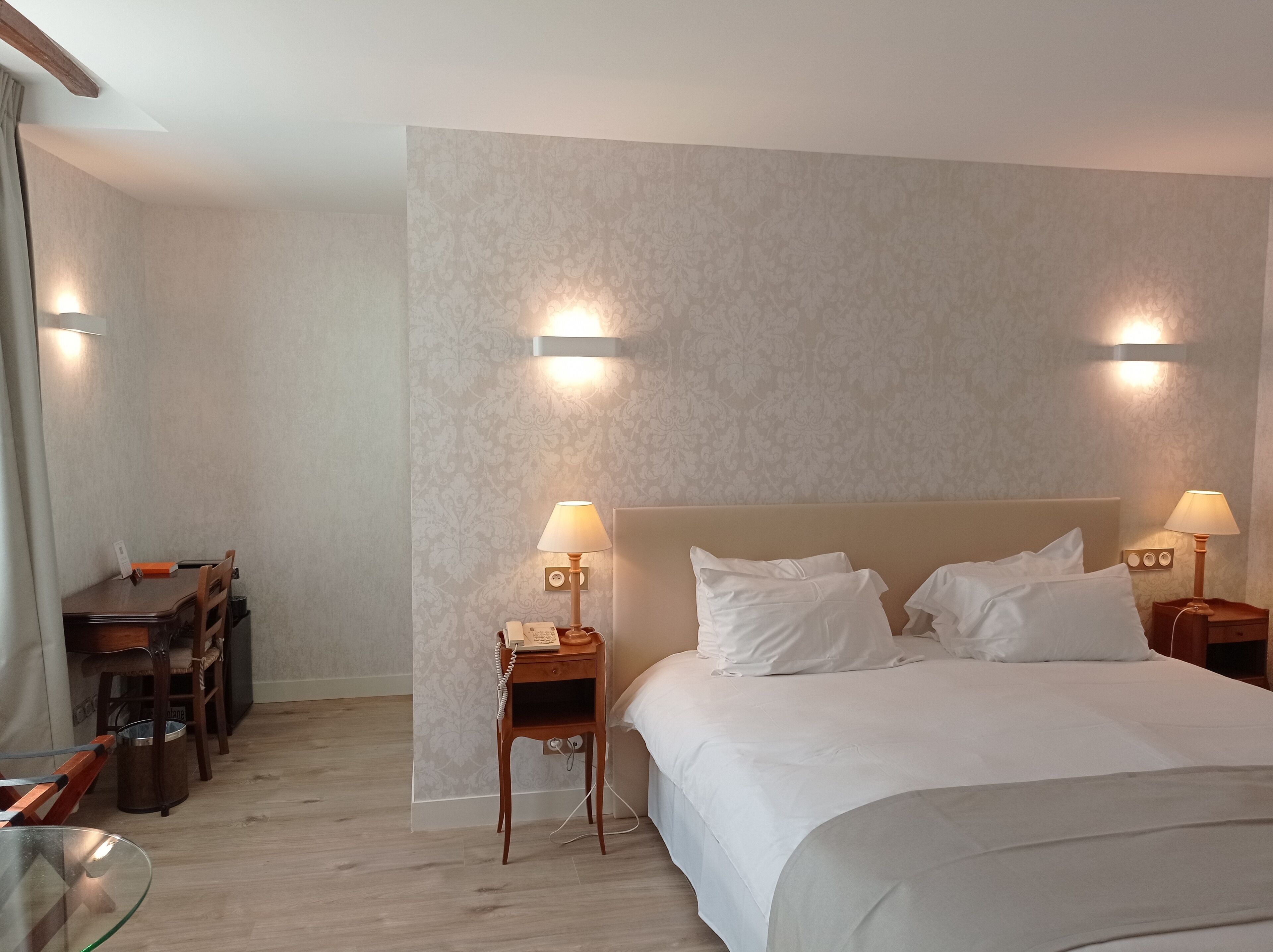 A hotel room with a large bed, two bedside lamps, a small table with a telephone, and a chair.