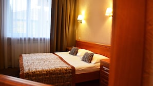 Una habitación de hotel con cama, mesita de noche, lámpara y ventana con cortinas.