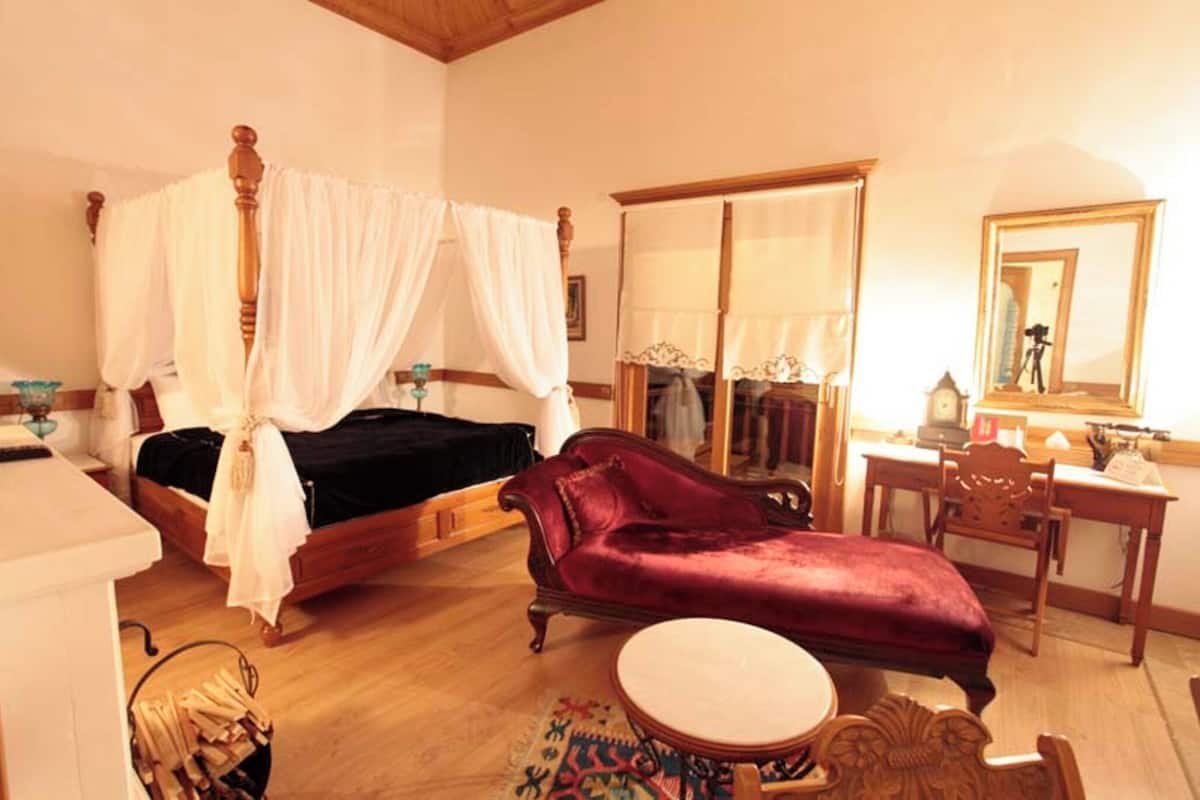 A bedroom with a four-poster bed, a red chaise lounge, a round table, and a mirror.