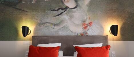 Habitación de hotel con un cuadro abstracto grande en la pared, cabecera gris, ropa de cama blanca y almohadas rojas.