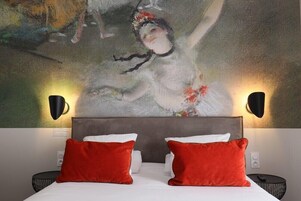 Habitación de hotel con un cuadro abstracto grande en la pared, cabecera gris, ropa de cama blanca y almohadas rojas.