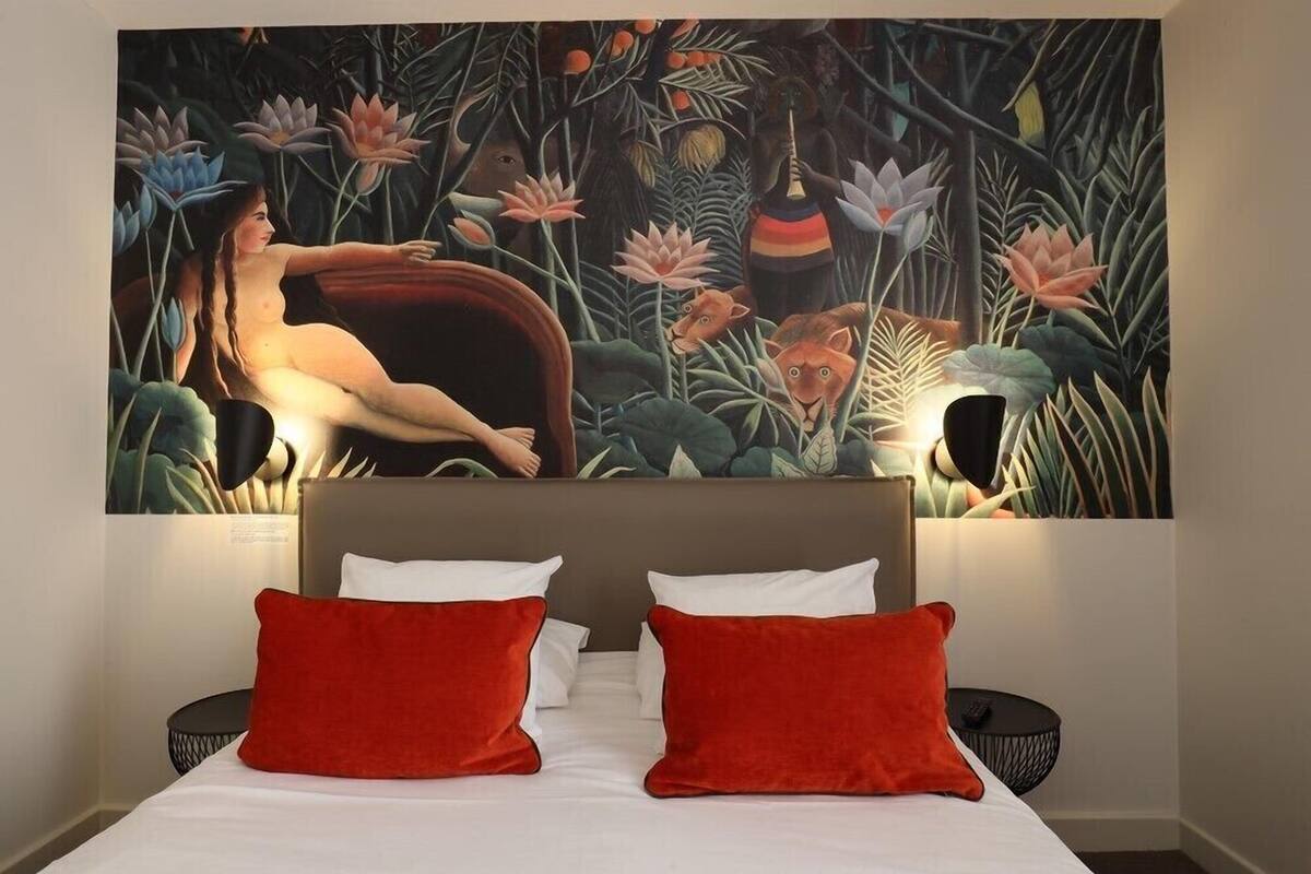 A hotel room with a large painting of a jungle scene above the bed, a bed with white and red pillows, and a round black side table.