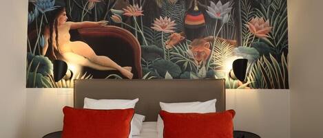 Quarto de hotel com uma grande pintura de uma cena de selva sobre a cama, uma cama com almofadas brancas e vermelhas e uma mesa lateral redonda preta.