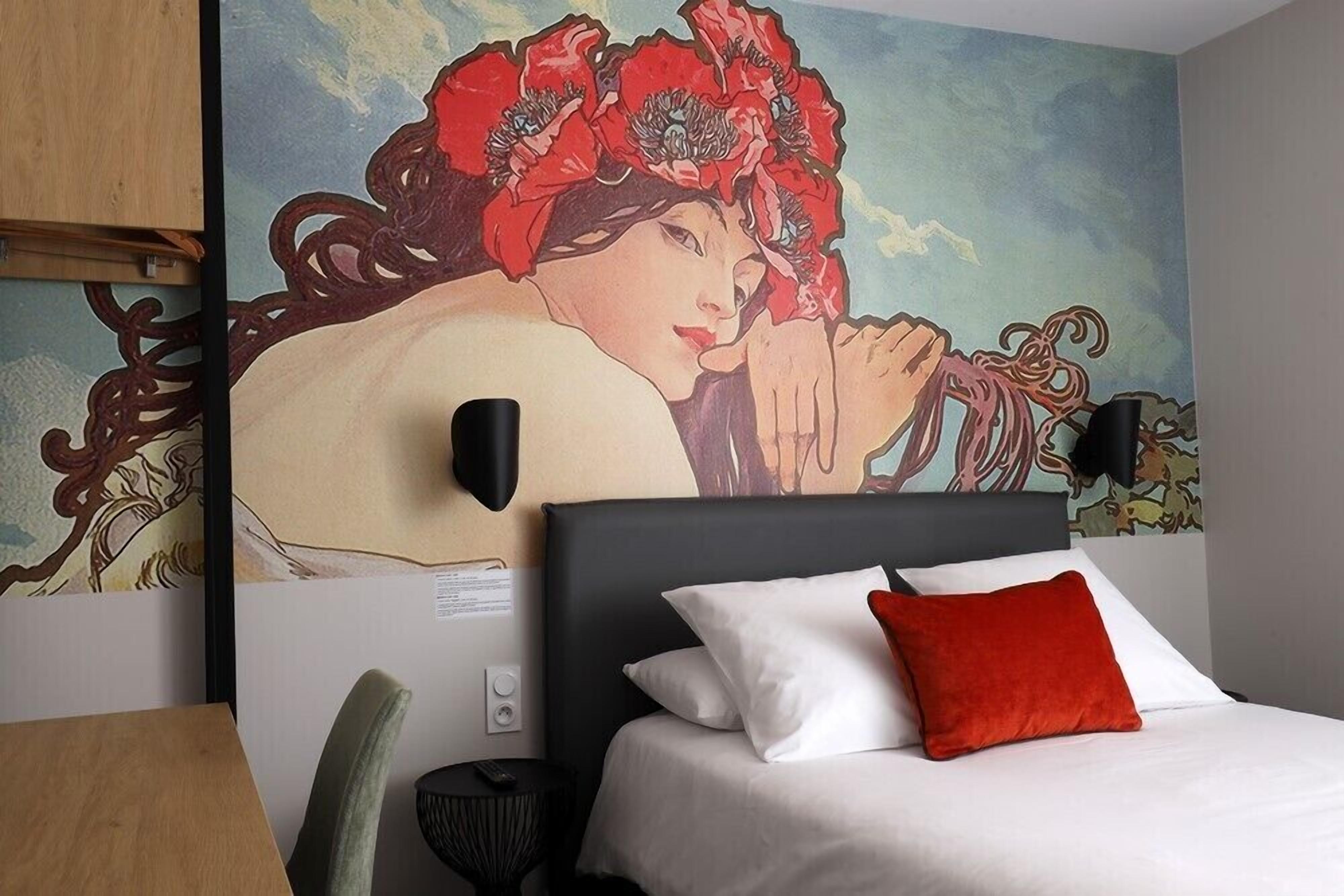 Una habitación de hotel con una cama, un almohánero rojo, una lámpara de pared y un mural grande.