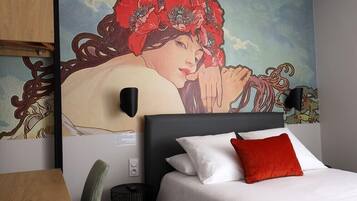 A hotel room with a bed, a red pillow, a wall-mounted lamp, and a large mural.