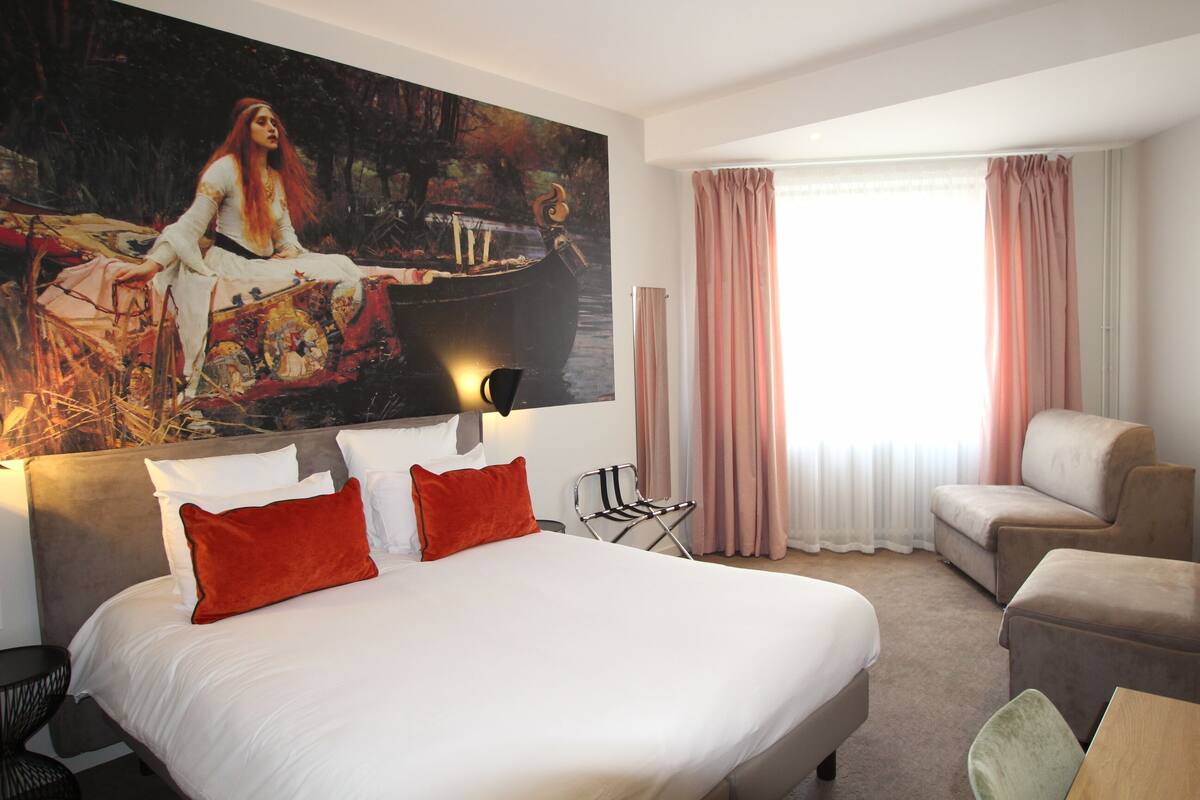 A hotel room with a large bed, a desk, a chair, and a painting on the wall.