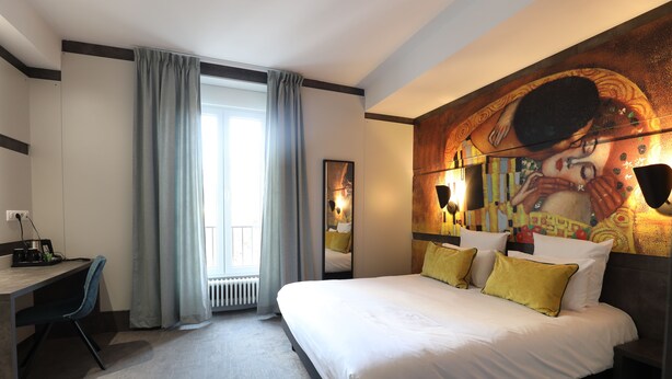 A hotel room with a large bed, a desk, and a chair. There is a window with curtains, a painting on the wall, and a radiator. - Hôtel  la Capelle (Millau)