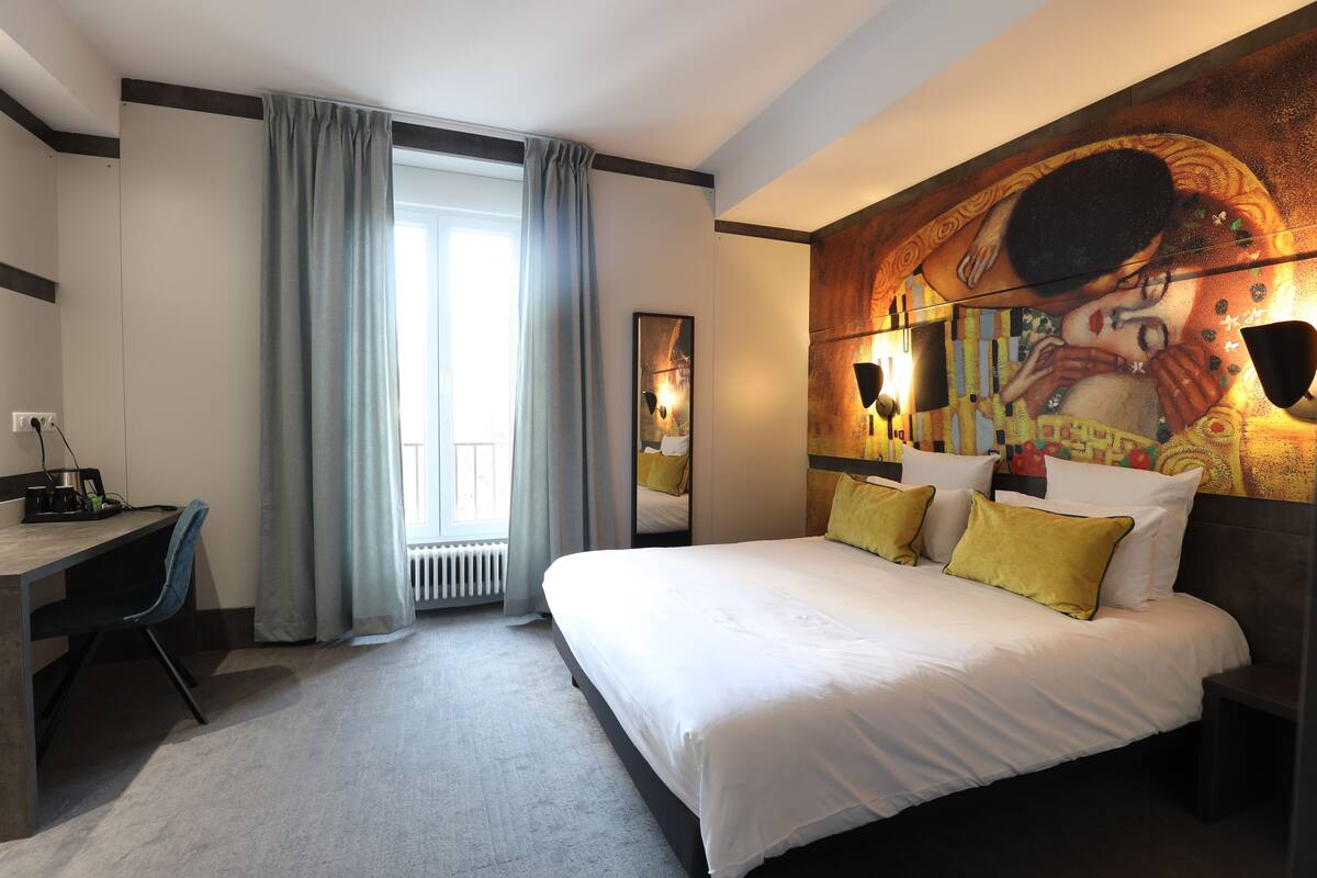 A hotel room with a large bed, a desk, and a chair. There is a window with curtains, a painting on the wall, and a radiator.