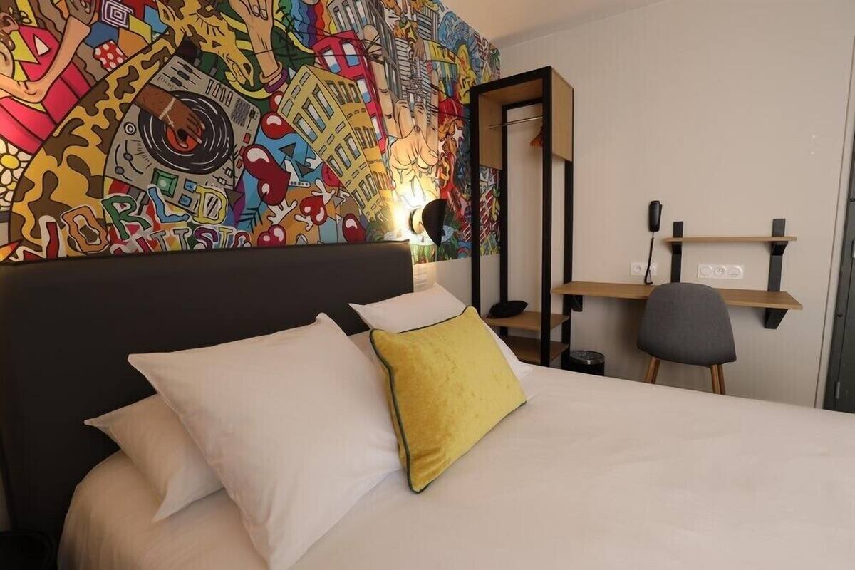 A hotel room with a bed, white and yellow pillows, a colorful abstract mural on the wall, a desk with a chair, and a shelf with a phone.