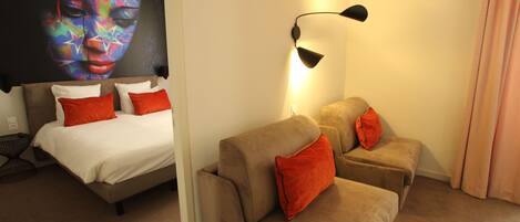 A hotel room with a bed, two sofas, a wall-mounted lamp, and a large abstract painting.